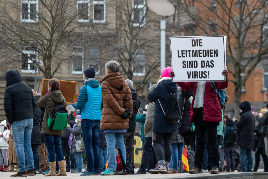 Frau mit Mundschutz hält ein Schild: Die Leidmedien sind das Virus!, dahinter Demo-Teilnehmer