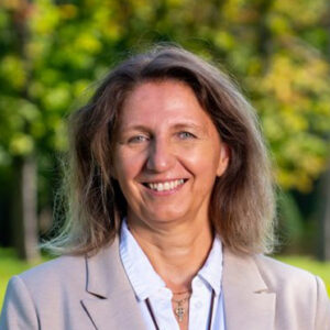 Frau lächelt im Freien, trägt Blazer und Bluse. Hintergrund mit unscharfen Bäumen.