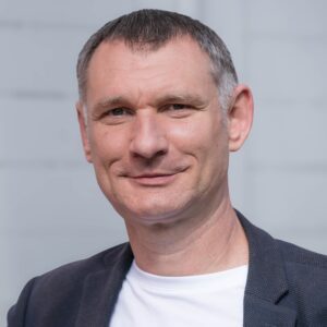 Mann mit kurzem Haar in Jacket und T-Shirt, lächelt leicht, neutrale Hintergrund.