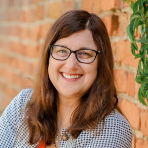 Frau mit Brille lächelt vor einer Ziegelwand.