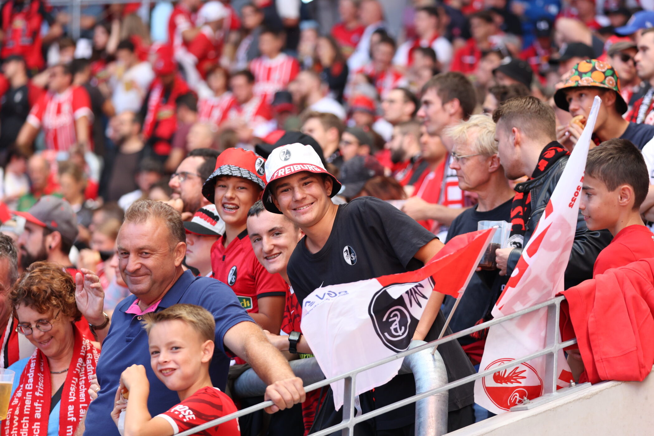 Menschenmenge in rot-weißer Kleidung, einige mit Hüten und Fahnen, lächelnd im Stadion.
