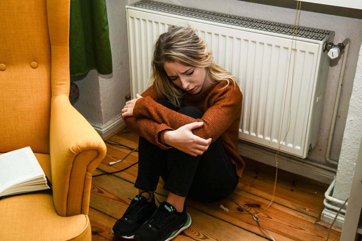 Person sitzt auf dem Boden vor einem Heizkörper, neben einem gelben Sessel.