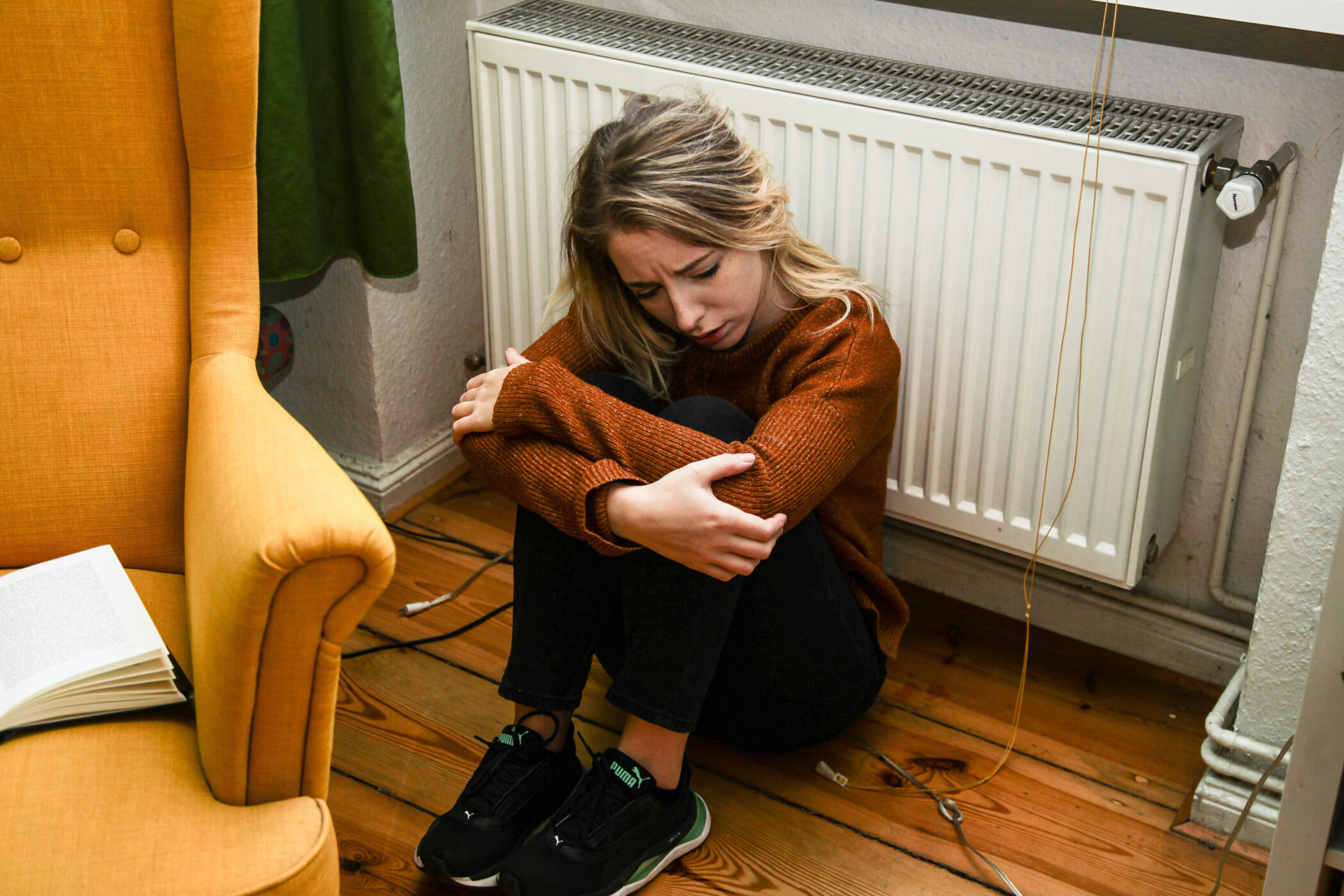 Person sitzt auf dem Boden vor einem Heizkörper, neben einem gelben Sessel.