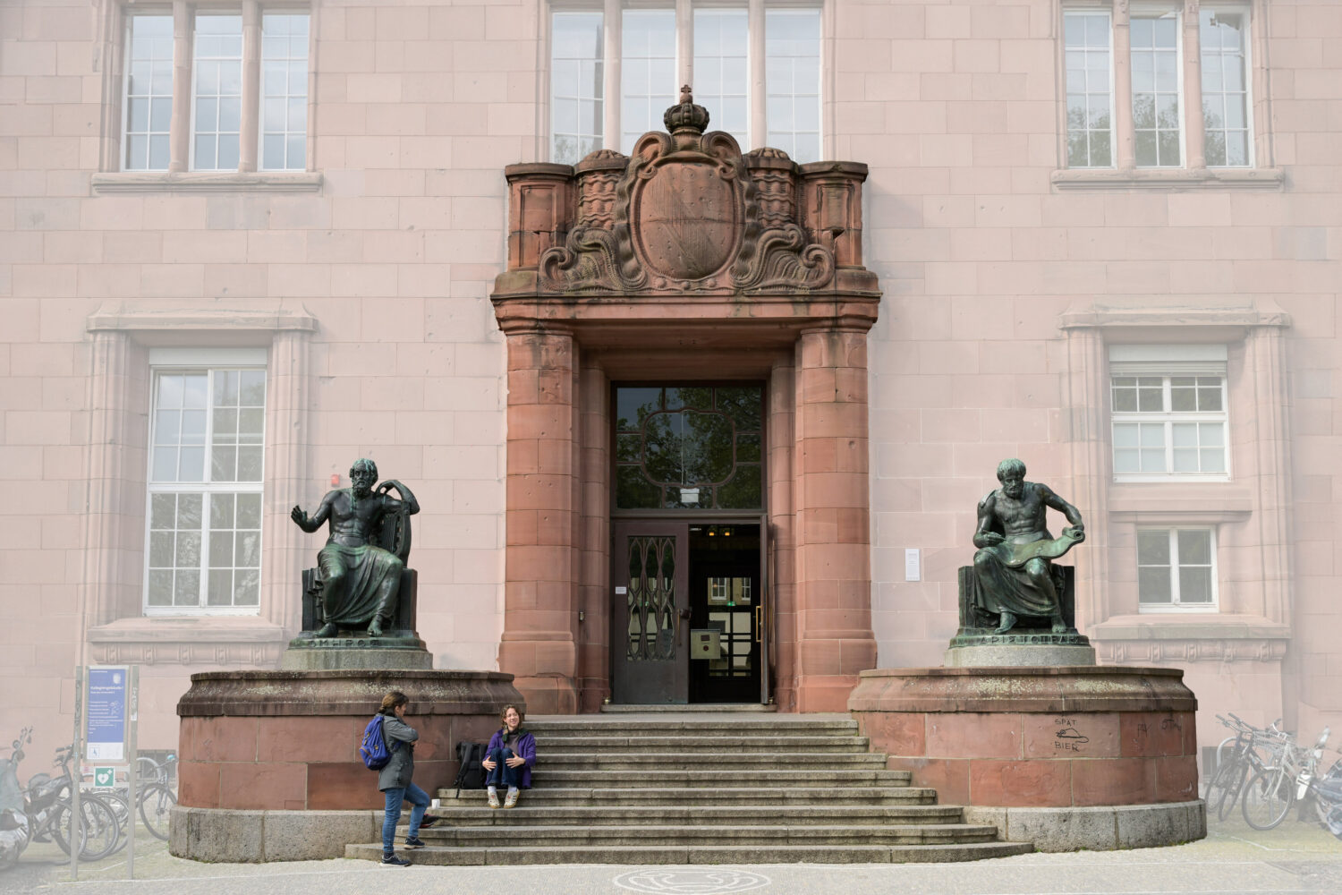Zwei Personen sitzen auf Treppe vor Gebäude mit zwei Statuen.