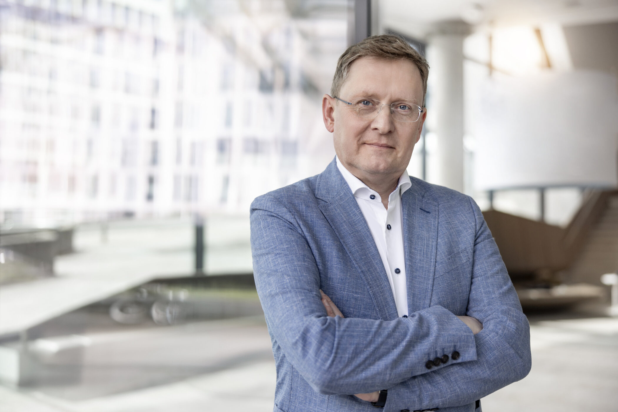 Mann in blauem Anzug mit verschränkten Armen vor Glasfassade.