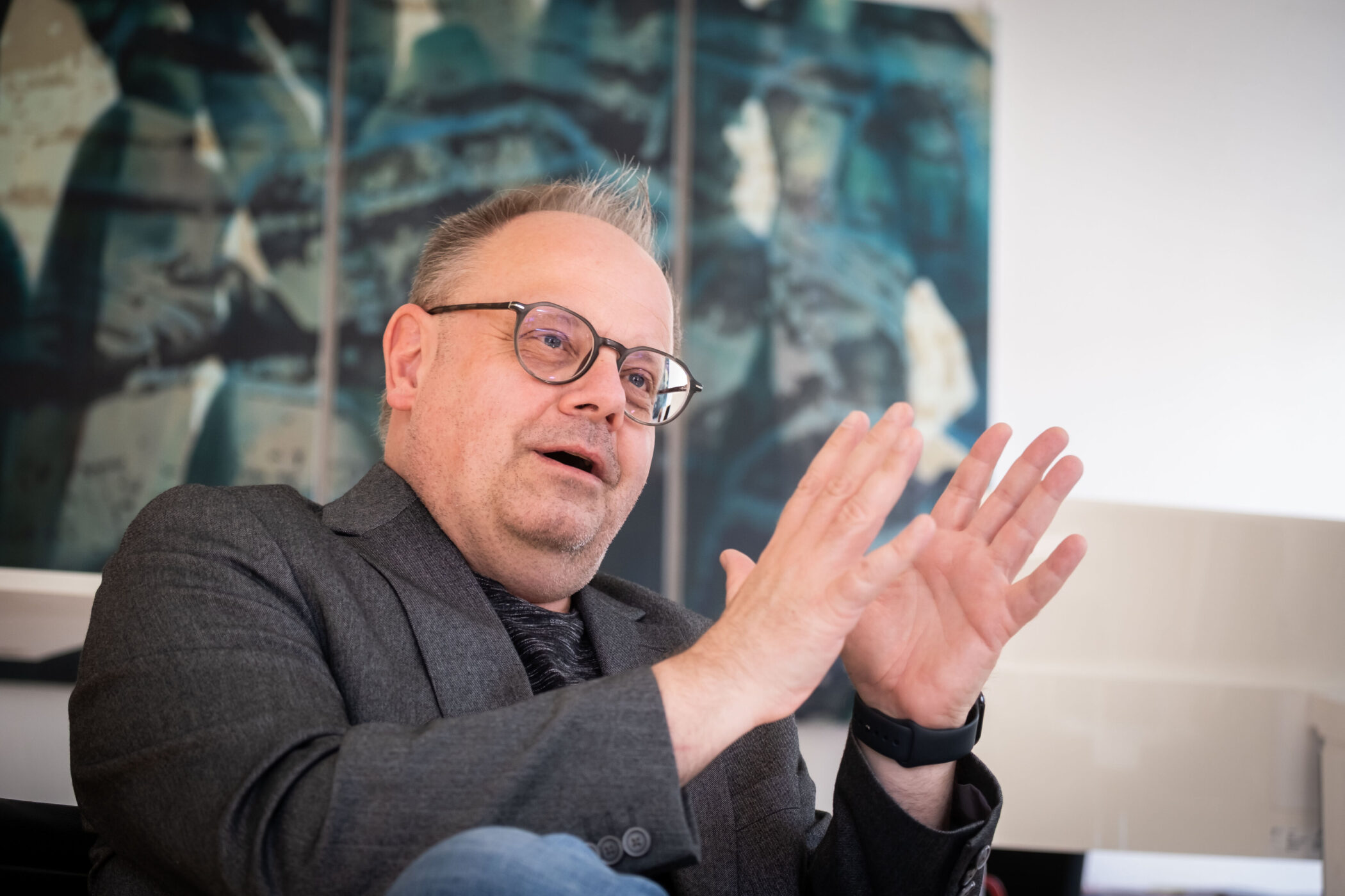 Mann mit Brille gestikuliert, im Hintergrund ein unscharfes Gemälde.