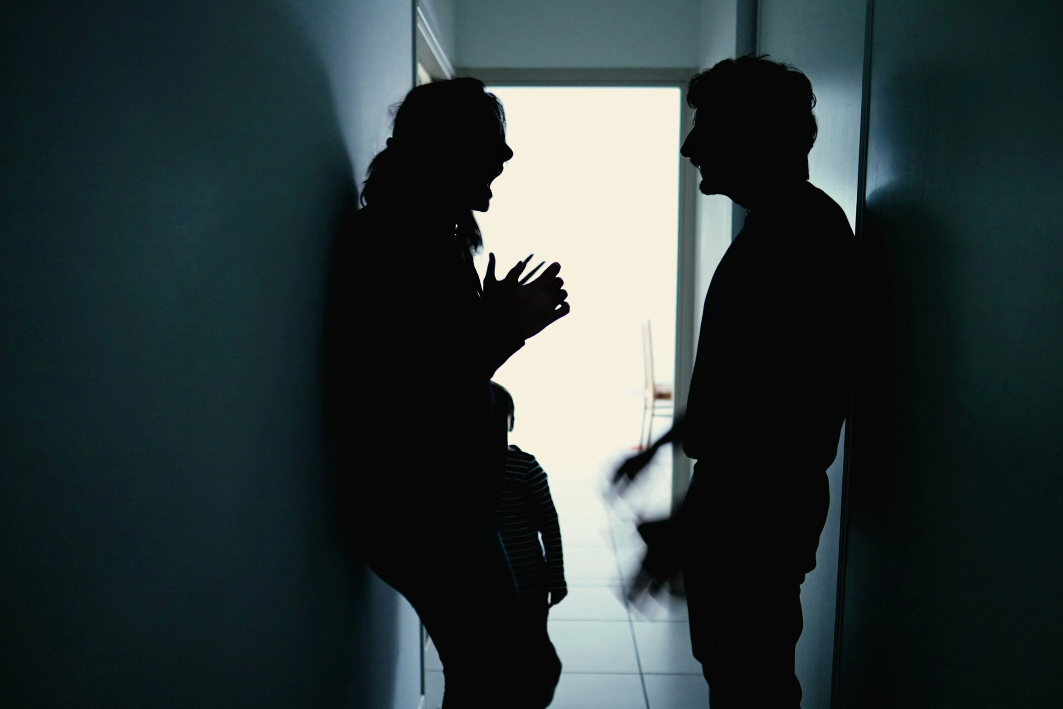 Zwei Personen stehen sich in einem dunklen Flur gegenüber, ein Kind im Hintergrund. Silhouetten vor hellem Türrahmen.