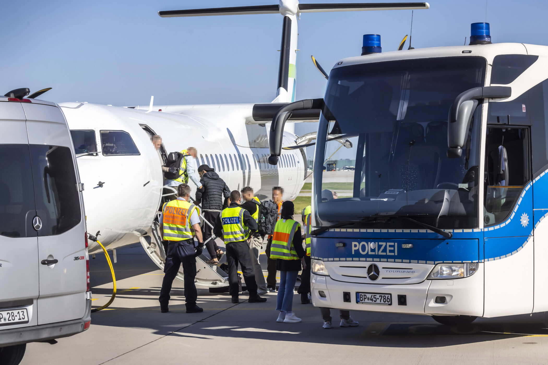 Personen steigen in ein Flugzeug, Polizei und Bus im Vordergrund.