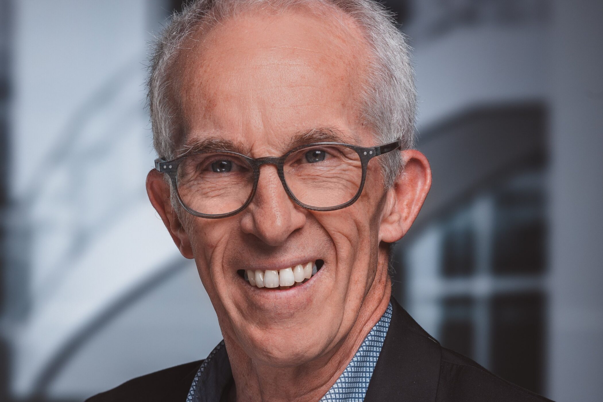 Älterer Mann mit Brille lächelt, trägt Anzug und Hemd, unscharfer Hintergrund.