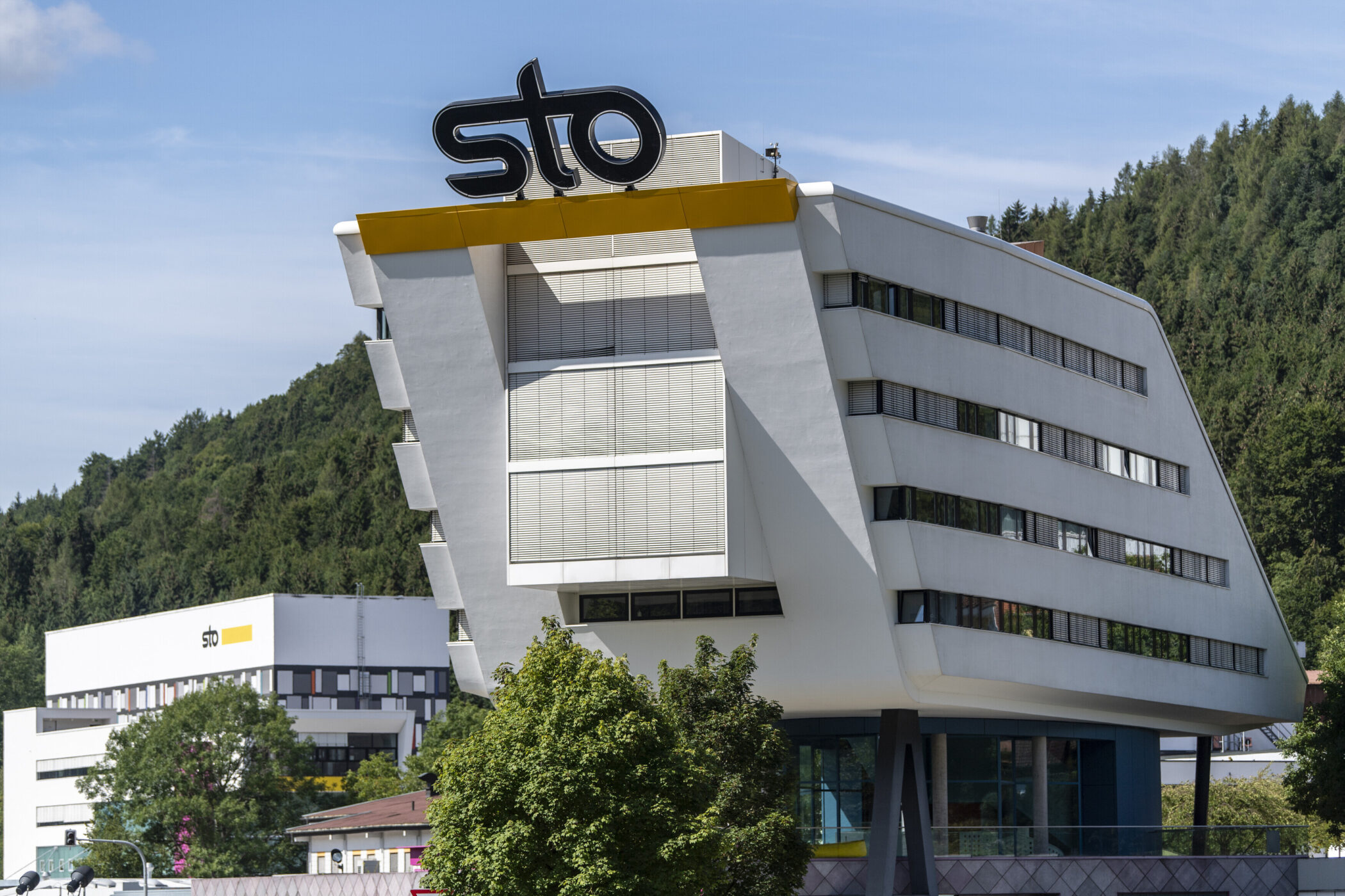 Modernes Gebäude mit "sto"-Logo, umgeben von Bäumen und weiteren Gebäuden.