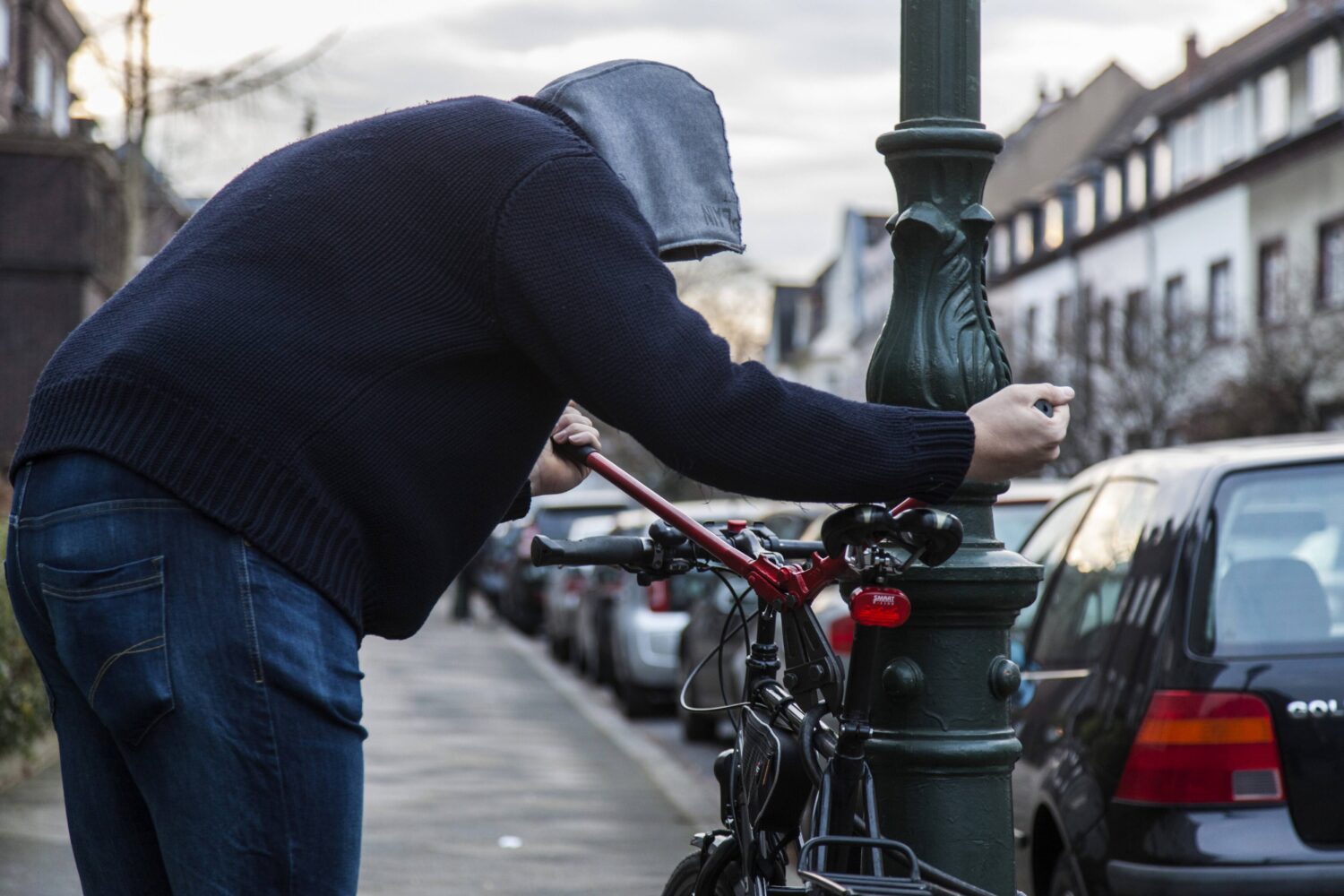 Person mit Kapuze beugt sich über ein Fahrrad an einem Laternenpfahl.