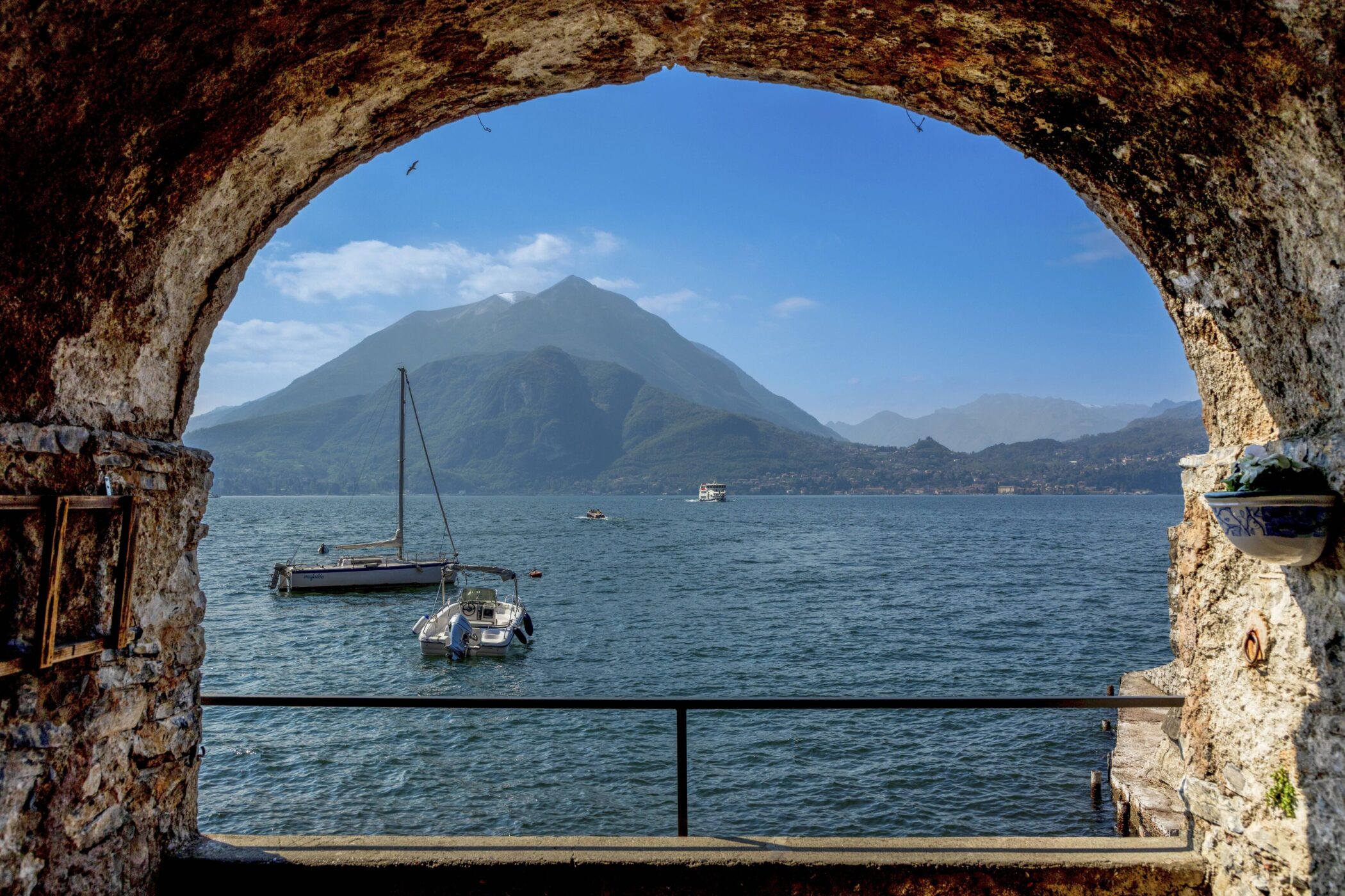 Blick durch einen Steinbogen auf einen See mit Booten und Bergen im Hintergrund.
