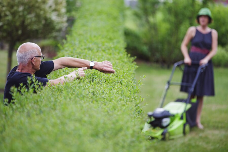 Mann zeigt auf Uhr, Frau im Hintergrund mit Rasenmäher.