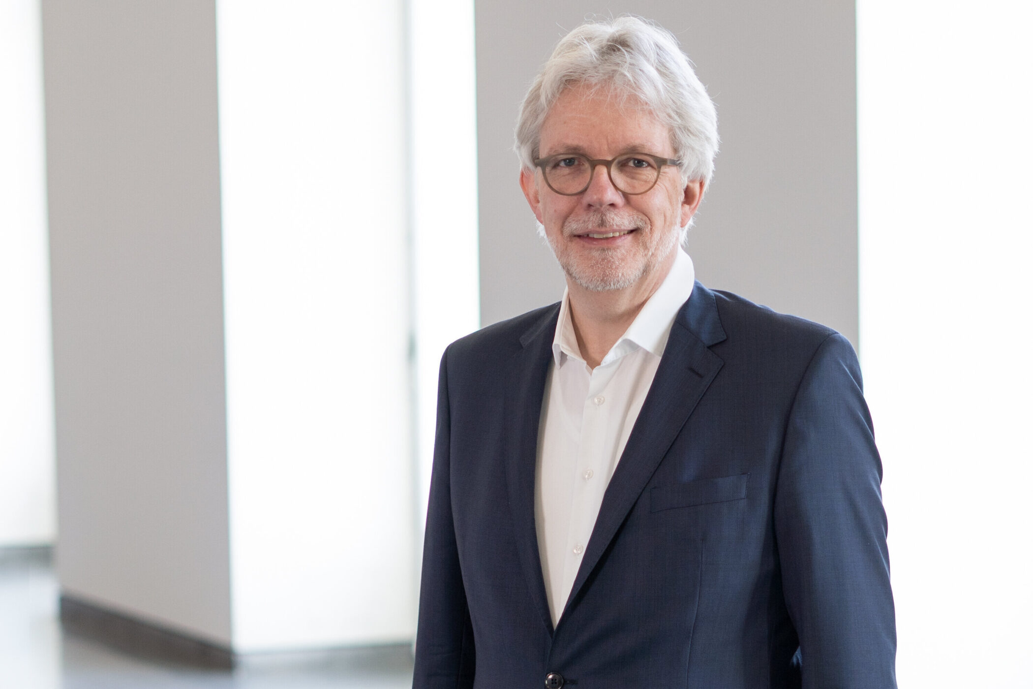 Älterer Mann mit Brille und Bart in Anzug, lächelt in heller Umgebung.