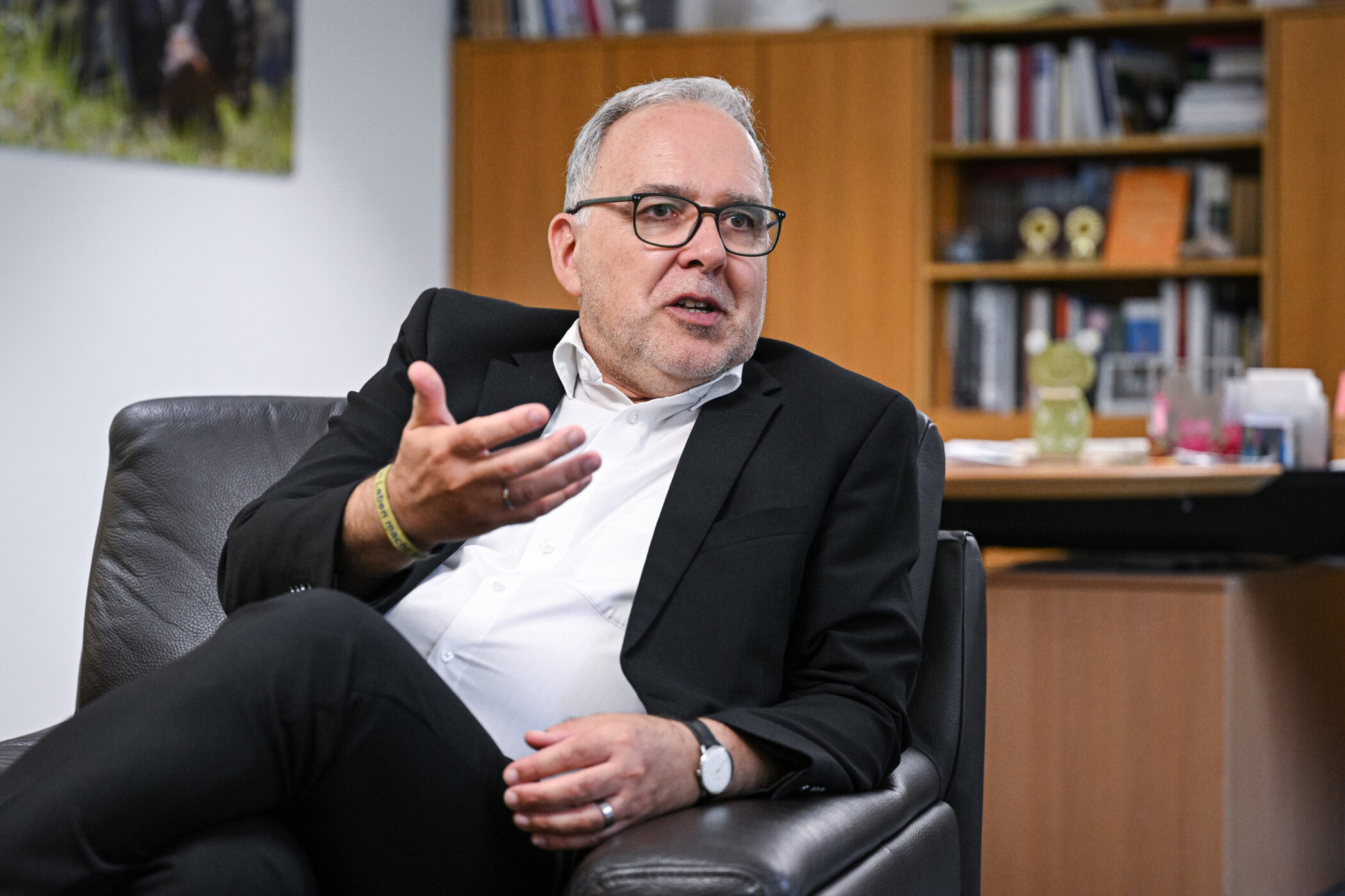 Mann im Anzug sitzt auf einem Ledersessel und gestikuliert. Bücherregal im Hintergrund.