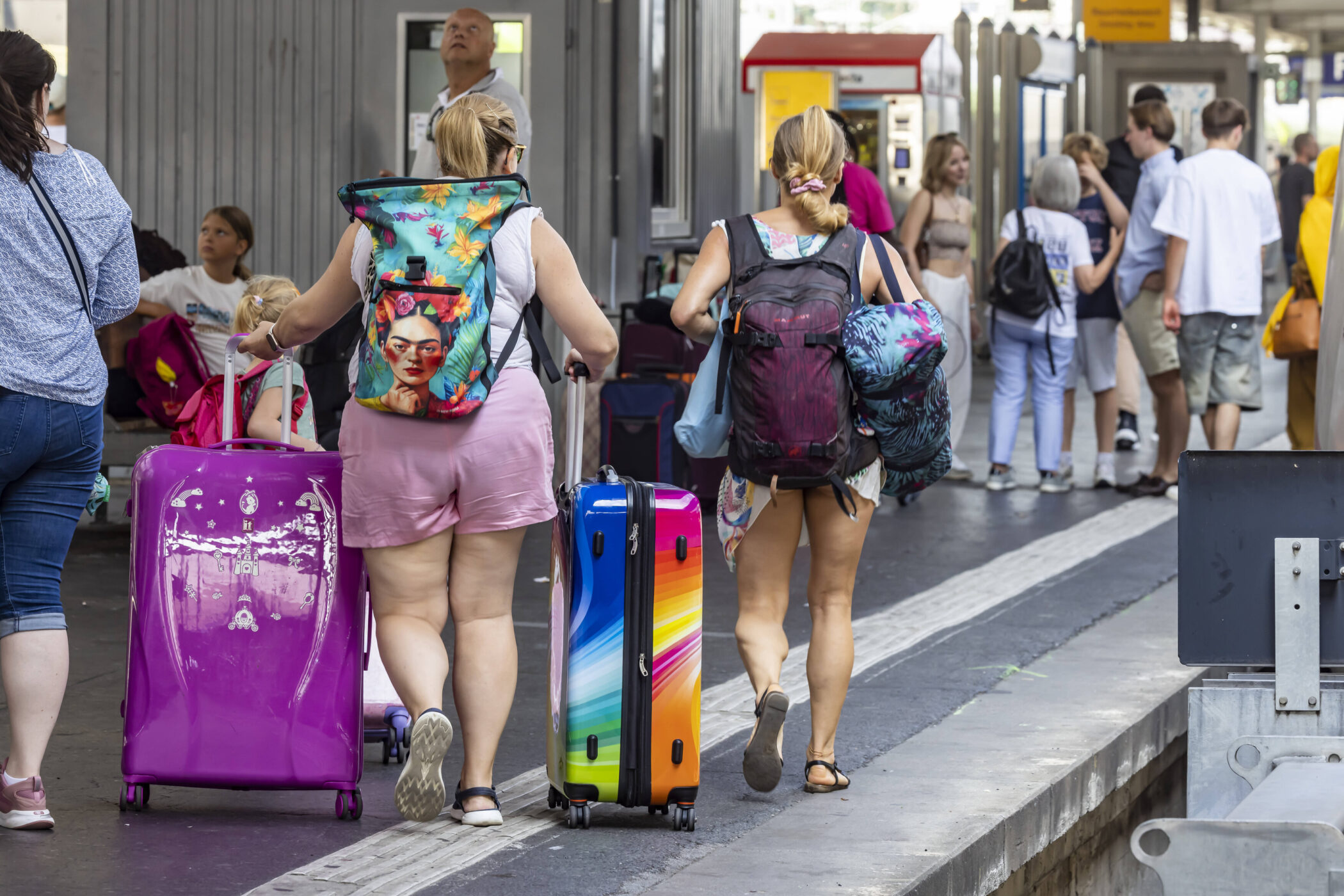 Zwei Personen mit bunten Koffern und Rucksäcken auf einem Bahnsteig.