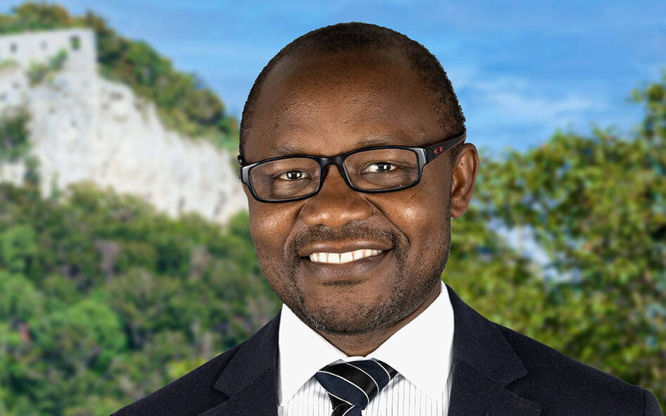 Mann mit Brille und Anzug lächelt vor unscharfem Naturhintergrund.