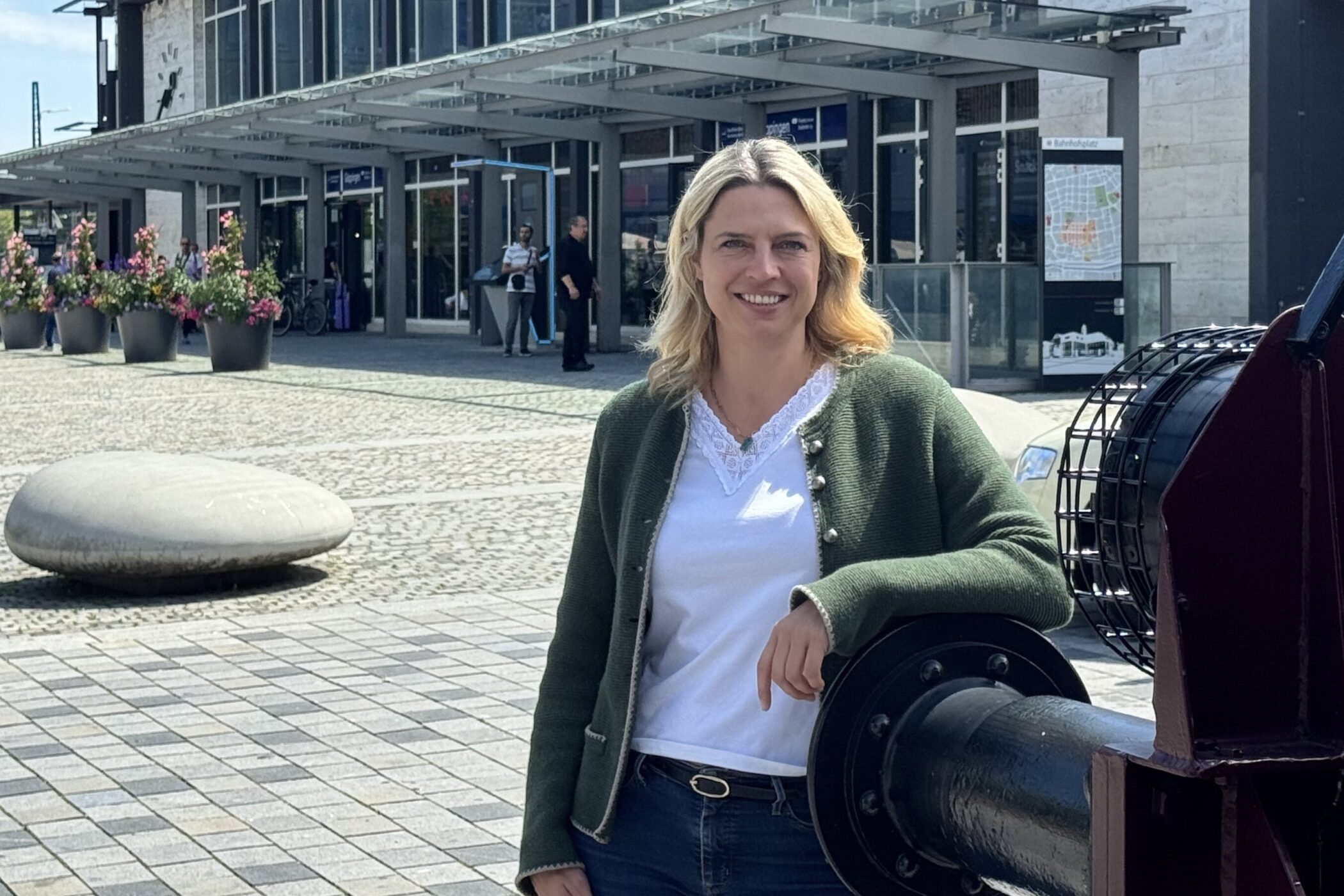 Eine Frau steht lächelnd vor einem modernen Gebäude mit Glasfassade.