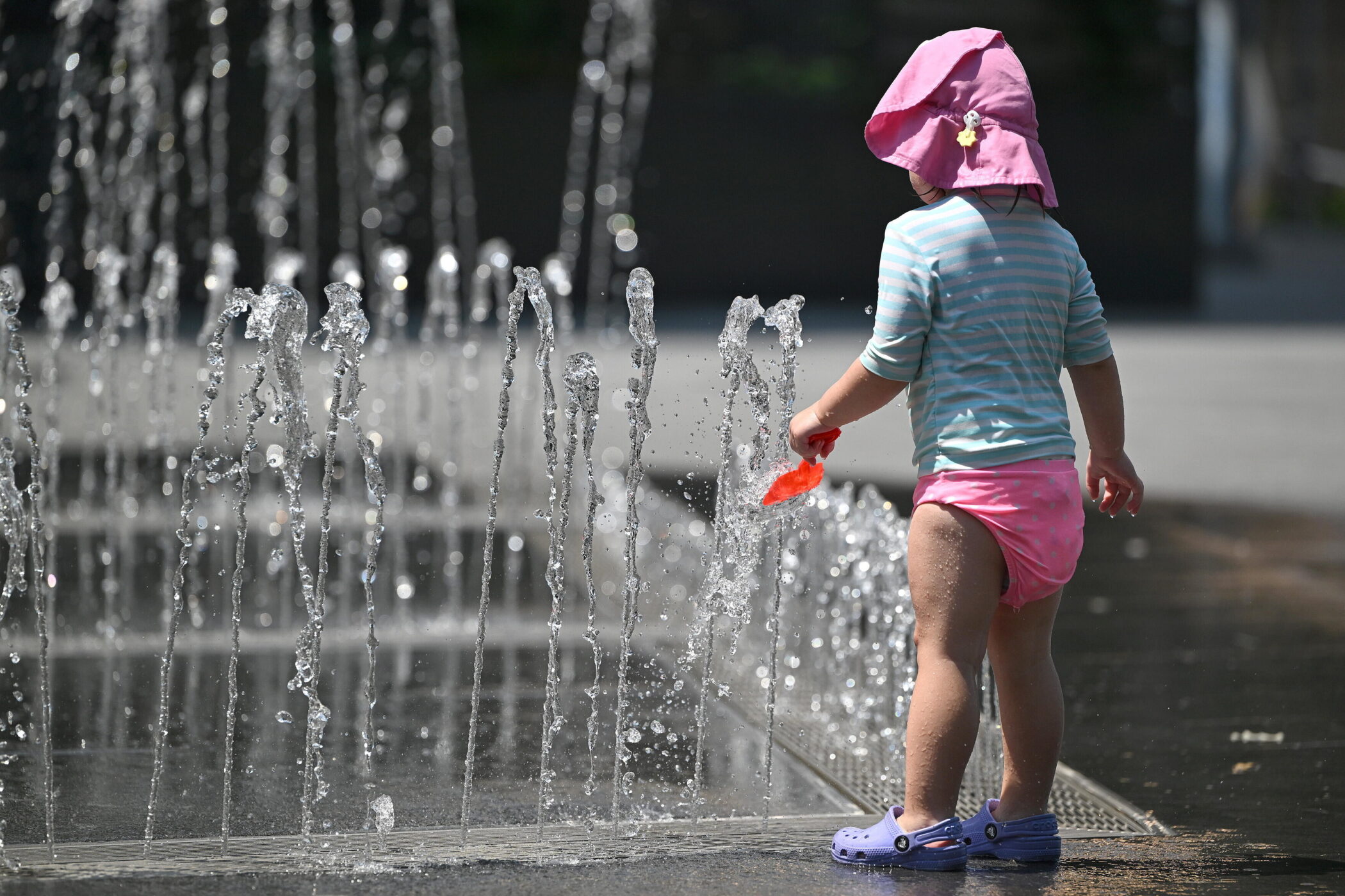 Kind mit pinkem Hut spielt an einem Springbrunnen.