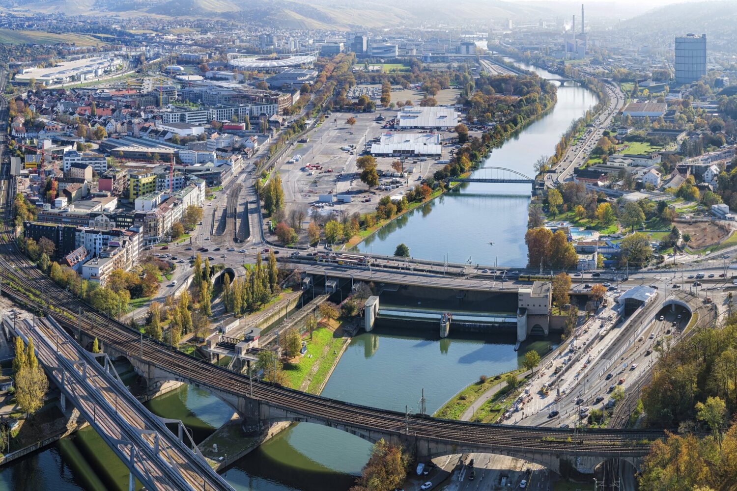 Stadtansicht mit Fluss, Brücken, Straßen und Gebäuden, umgeben von Hügeln und Bäumen.