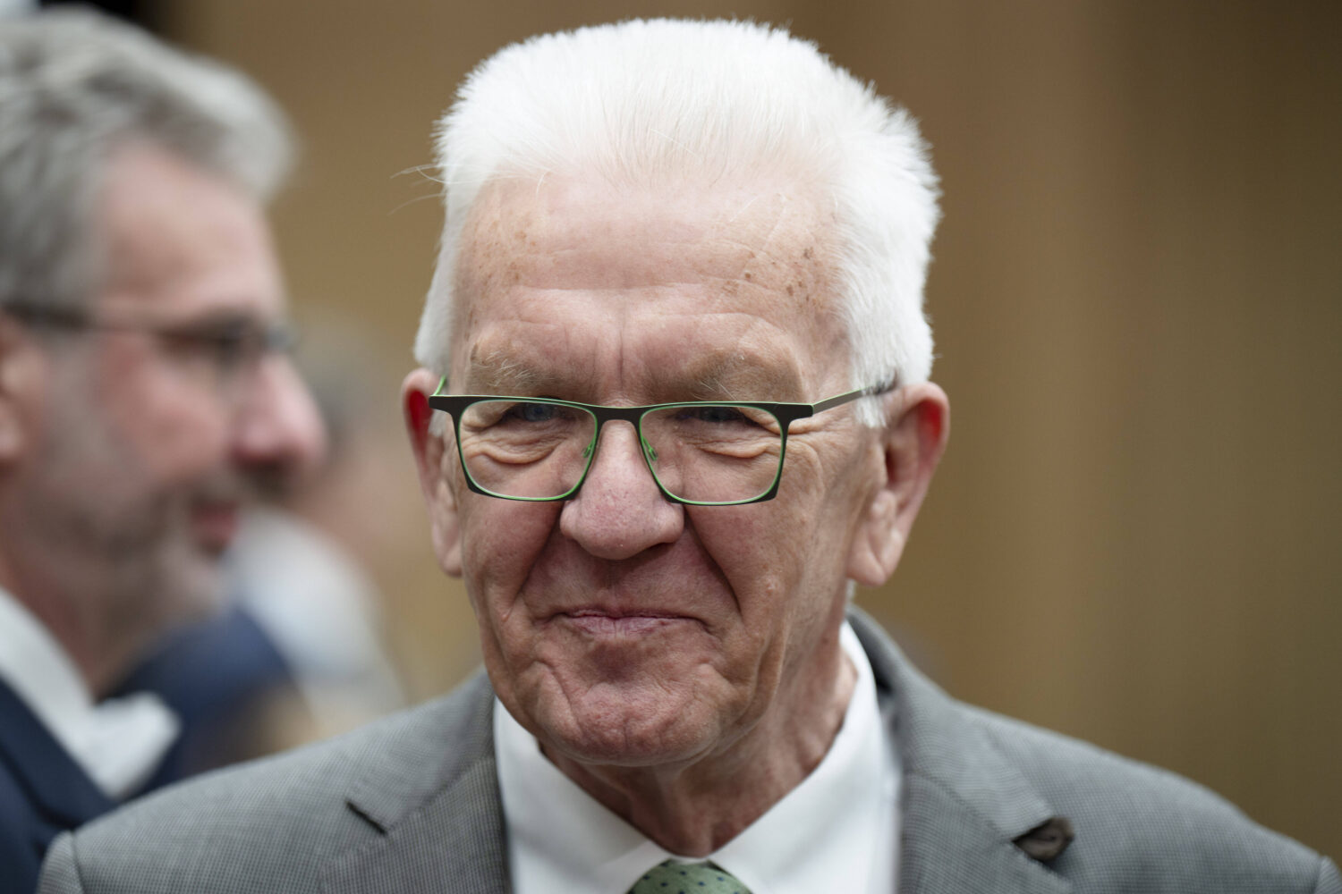 Älterer Mann mit Brille und grauem Anzug, lächelnd, unscharfe Person im Hintergrund.