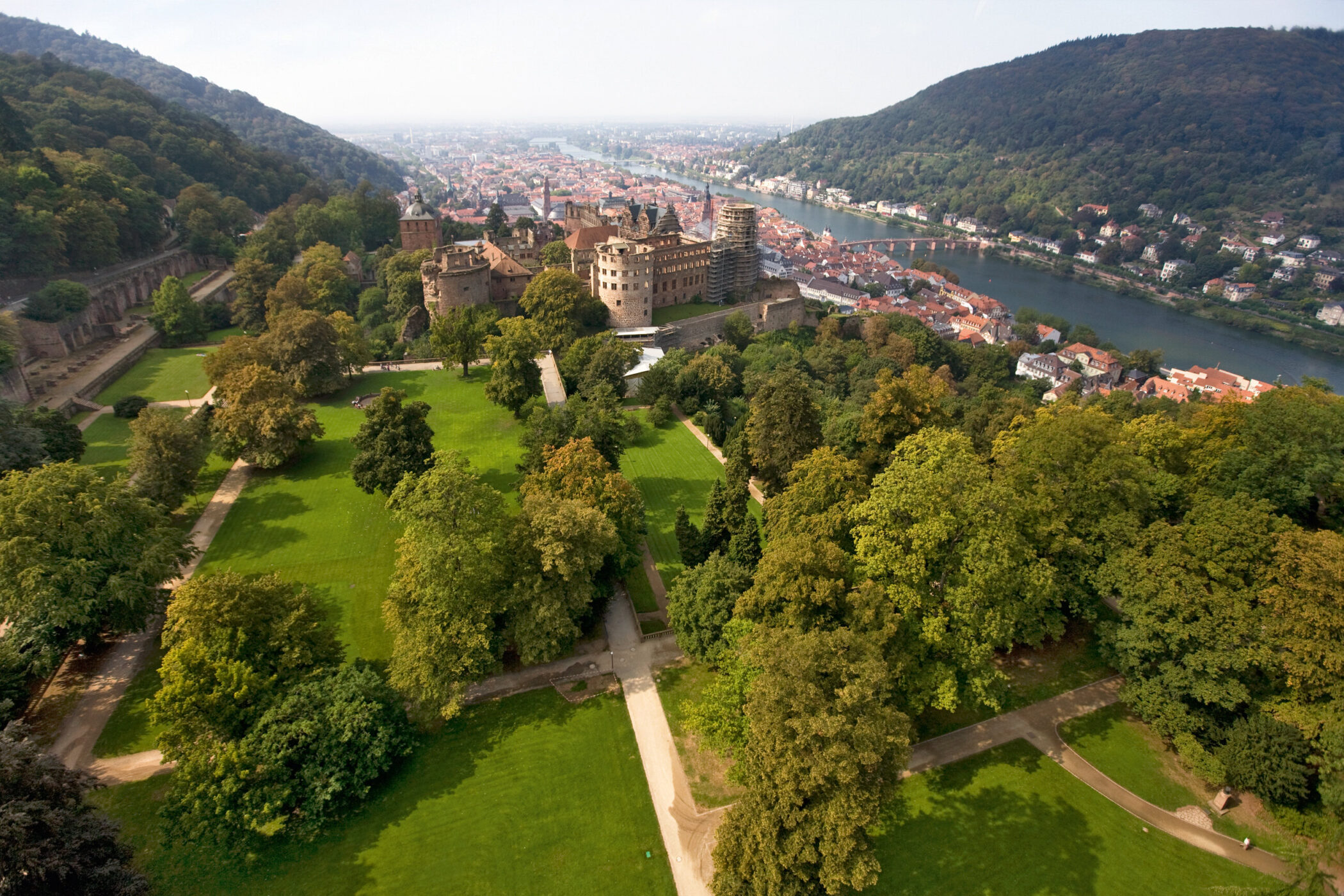 Schloss mit Gartenanlage, Fluss und Stadt im Hintergrund, umgeben von bewaldeten Hügeln.