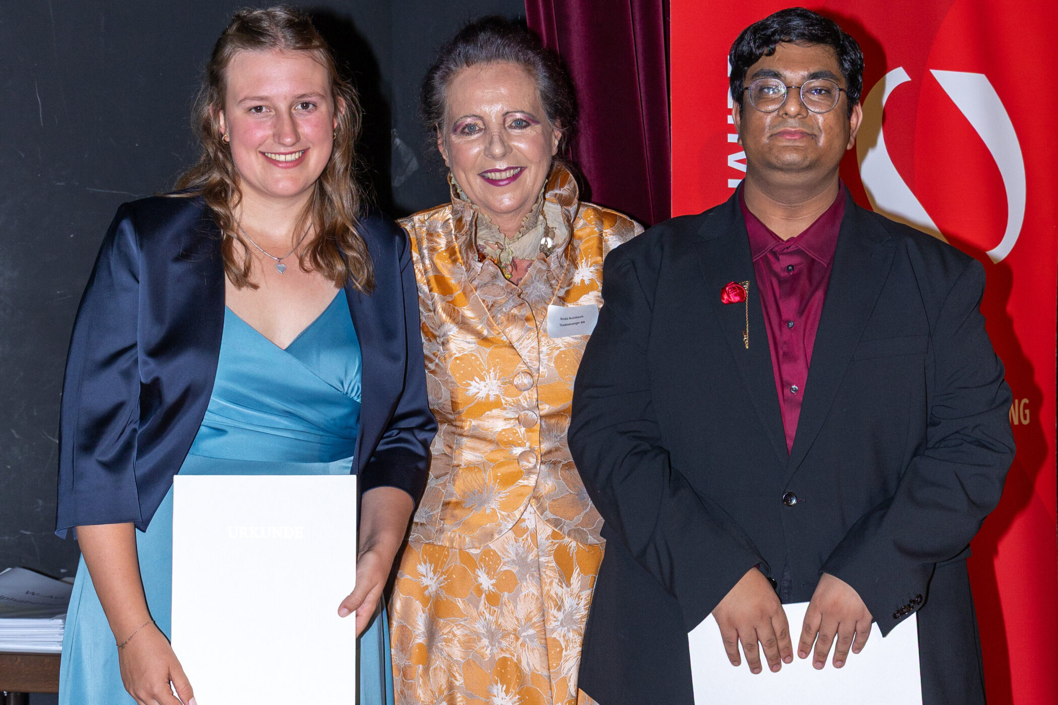 Drei Personen posieren, zwei halten Urkunden, vor rotem Hintergrund.