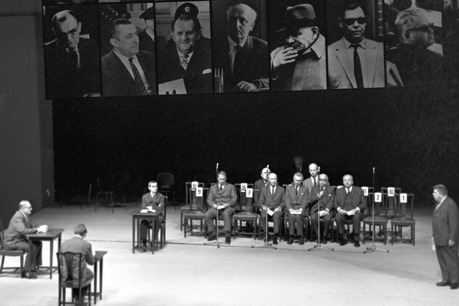 Mehrere Männer in Anzügen sitzen auf einer Bühne, einige stehen. Oben sind Porträts.