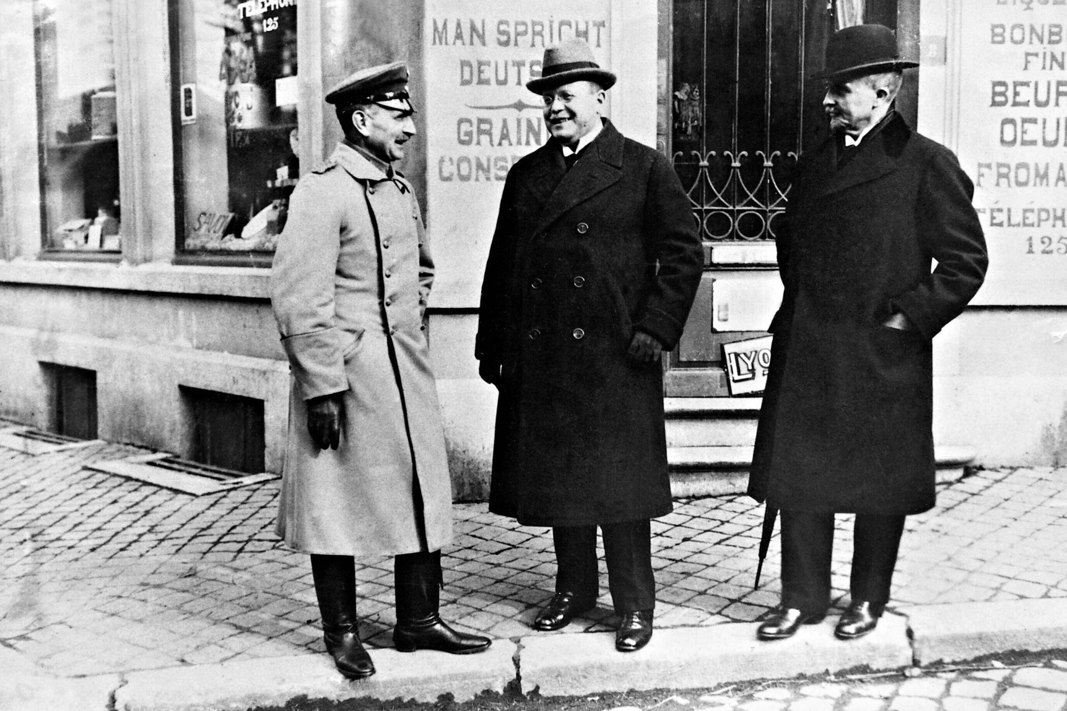 Drei Männer in Mänteln und Hüten stehen auf einer gepflasterten Straße und unterhalten sich.