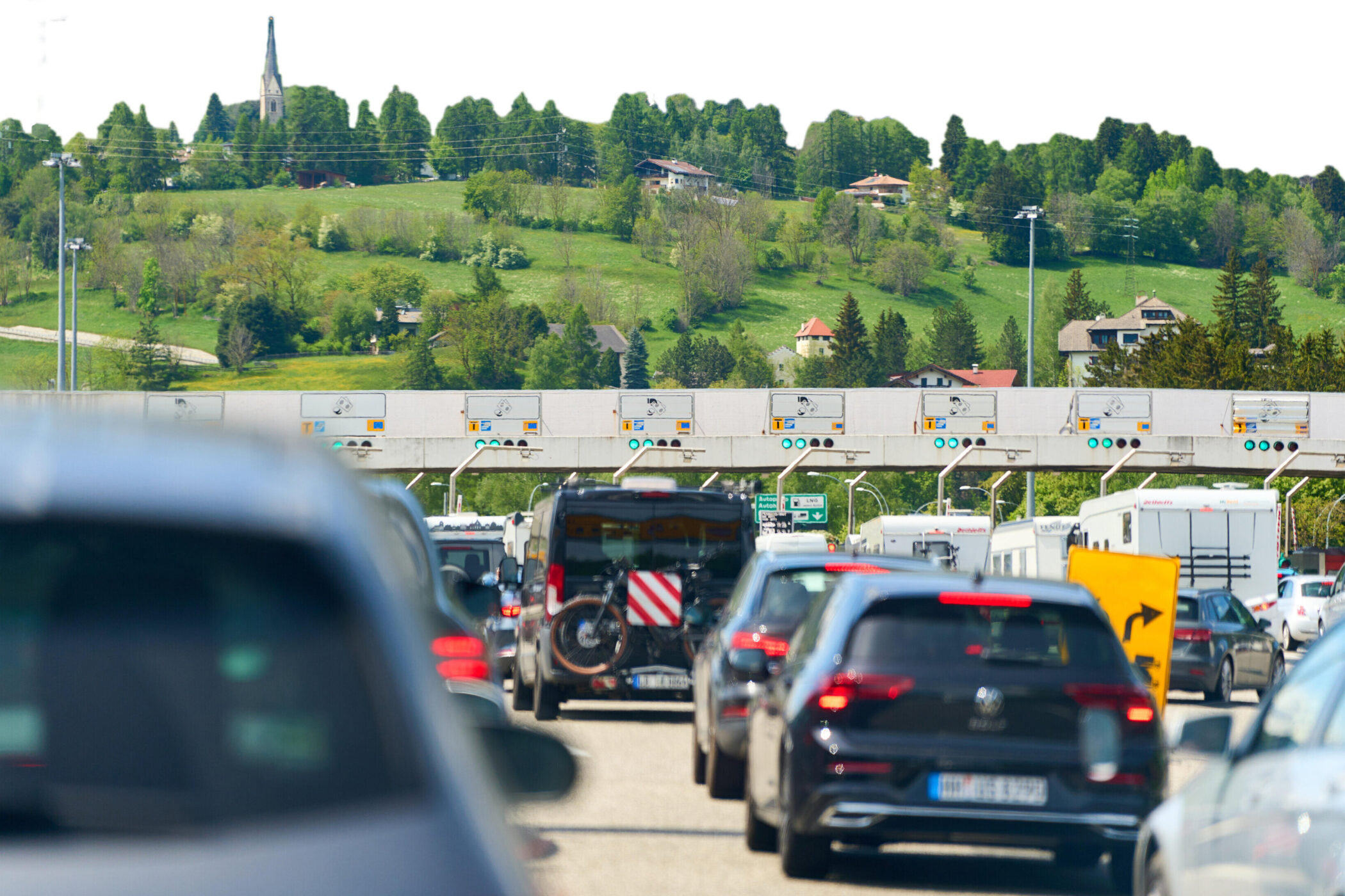 Autoschlange vor Mautstelle, im Hintergrund grüne Hügel und Gebäude.
