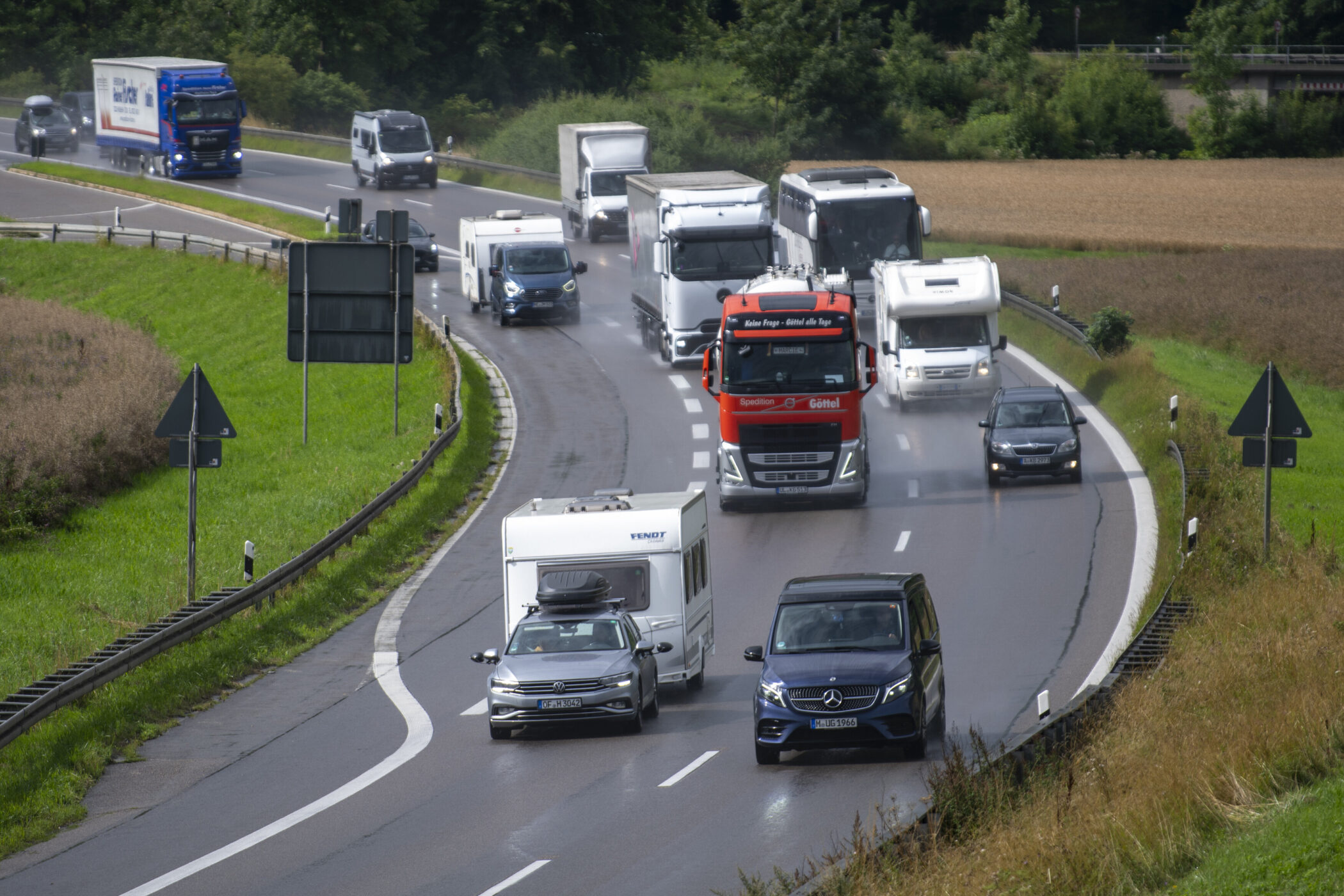Mehrere Fahrzeuge auf einer nassen Landstraße, darunter Lkw und Wohnmobile.