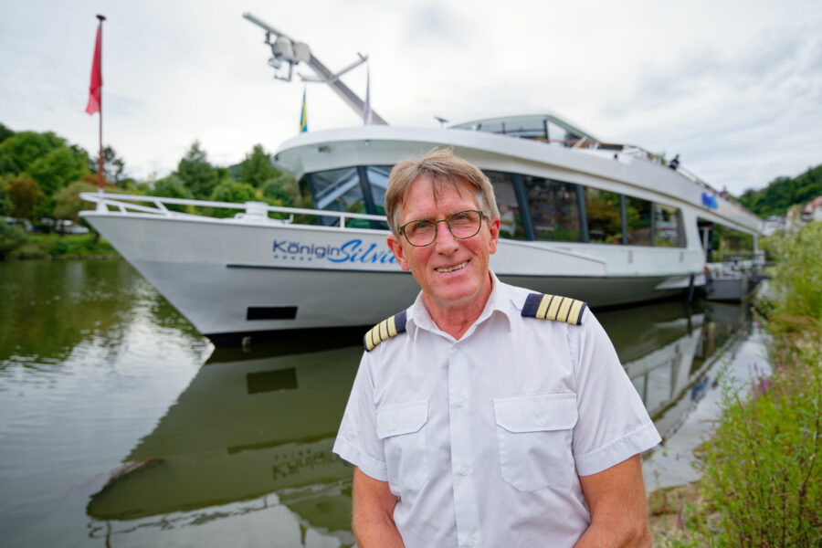 Mann in Uniform vor einem weißen Schiff mit der Aufschrift "Königin Silvia" am Fluss.