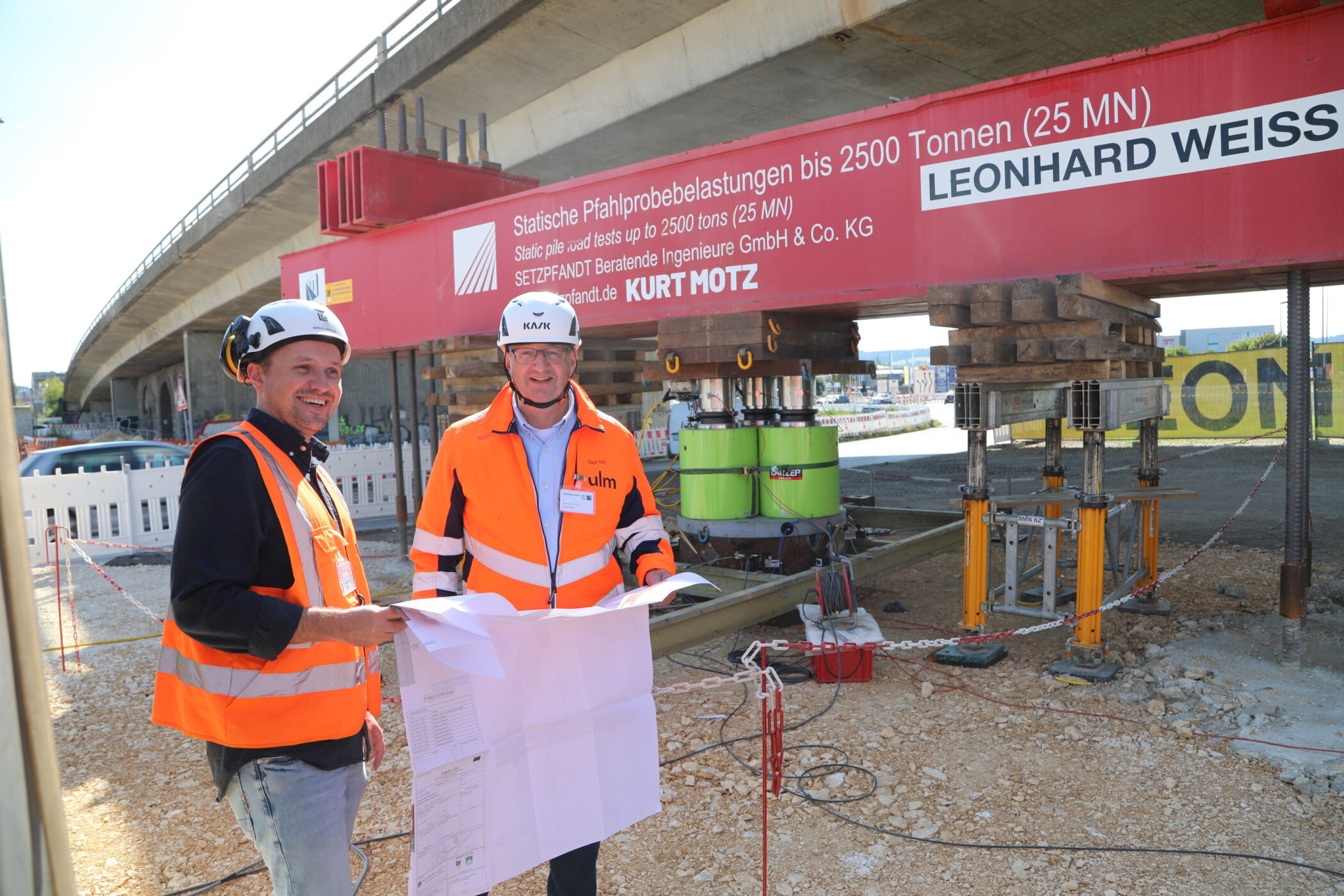 Zwei Männer in Warnwesten und Helmen, halten Baupläne unter einer Brücke.