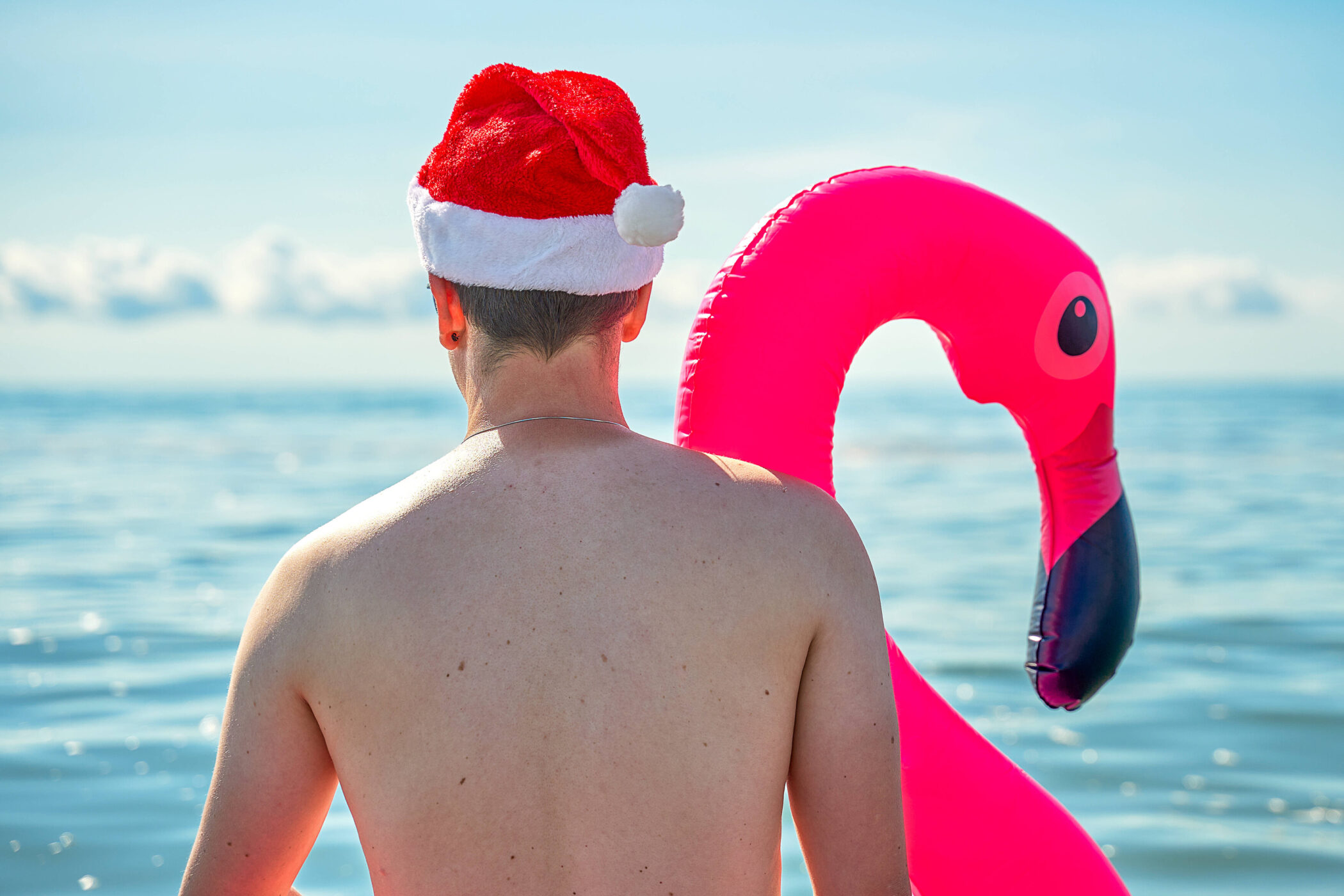 Person mit Weihnachtsmütze und rosa Flamingo-Schwimmring am Meer.