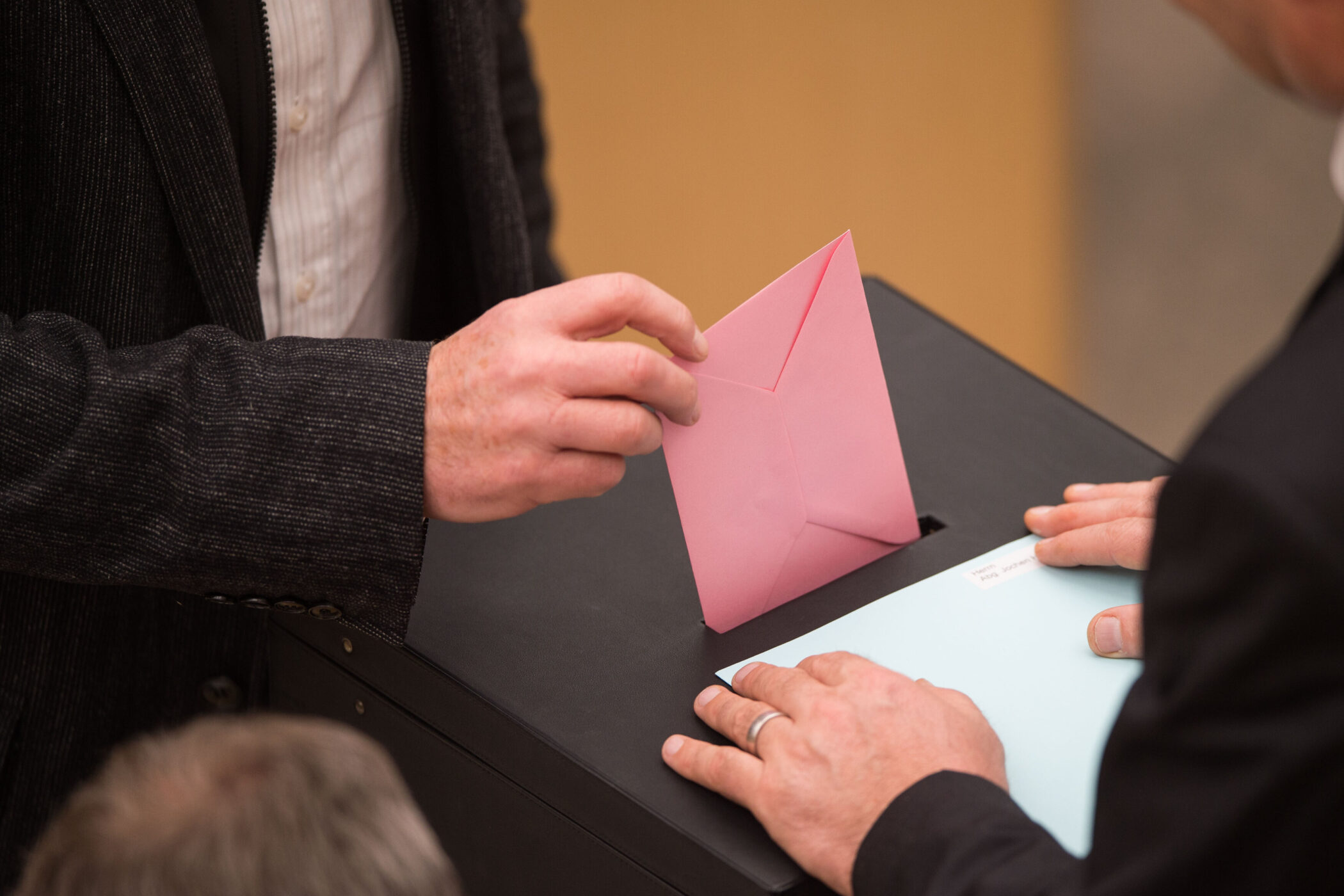 Person steckt rosa Umschlag in eine schwarze Box, andere Person hält blauen Umschlag.