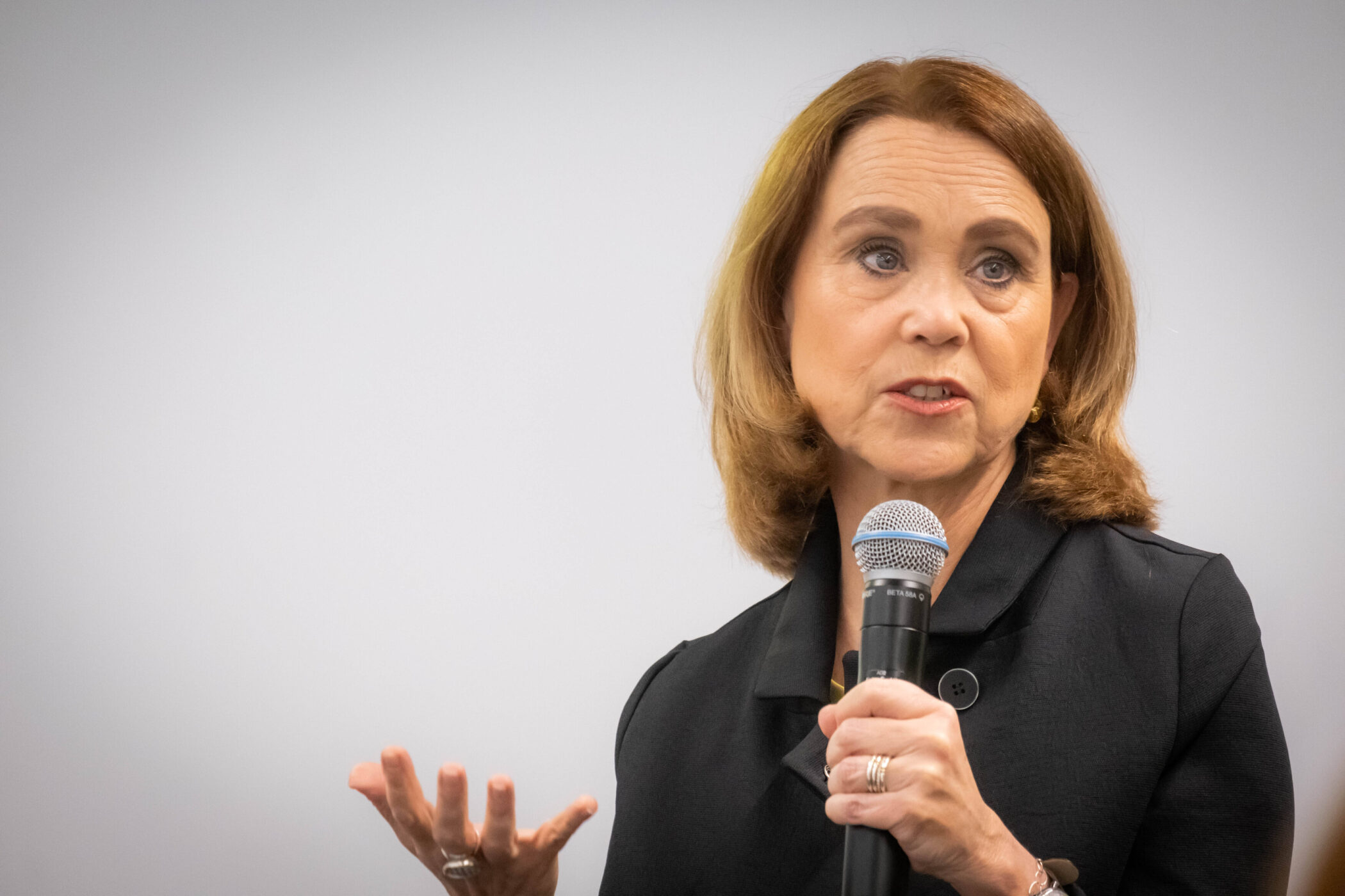 Frau mit Mikrofon spricht, hebt eine Hand, trägt schwarze Kleidung.