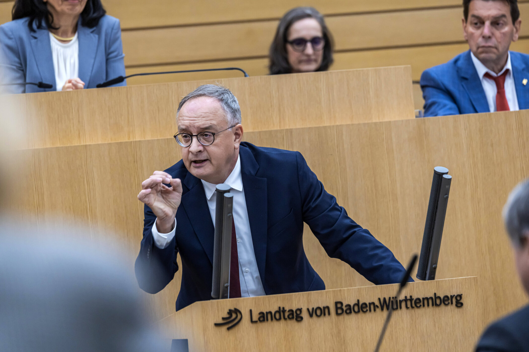 Mann in Anzug spricht im Landtag von Baden-Württemberg, drei Personen im Hintergrund.