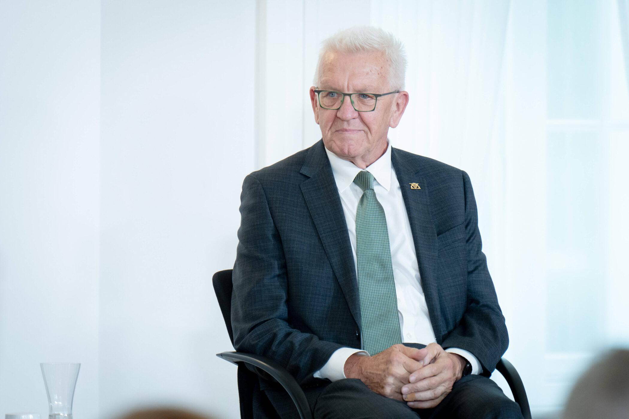 Älterer Mann in Anzug sitzt auf Stuhl, schaut zur Seite.