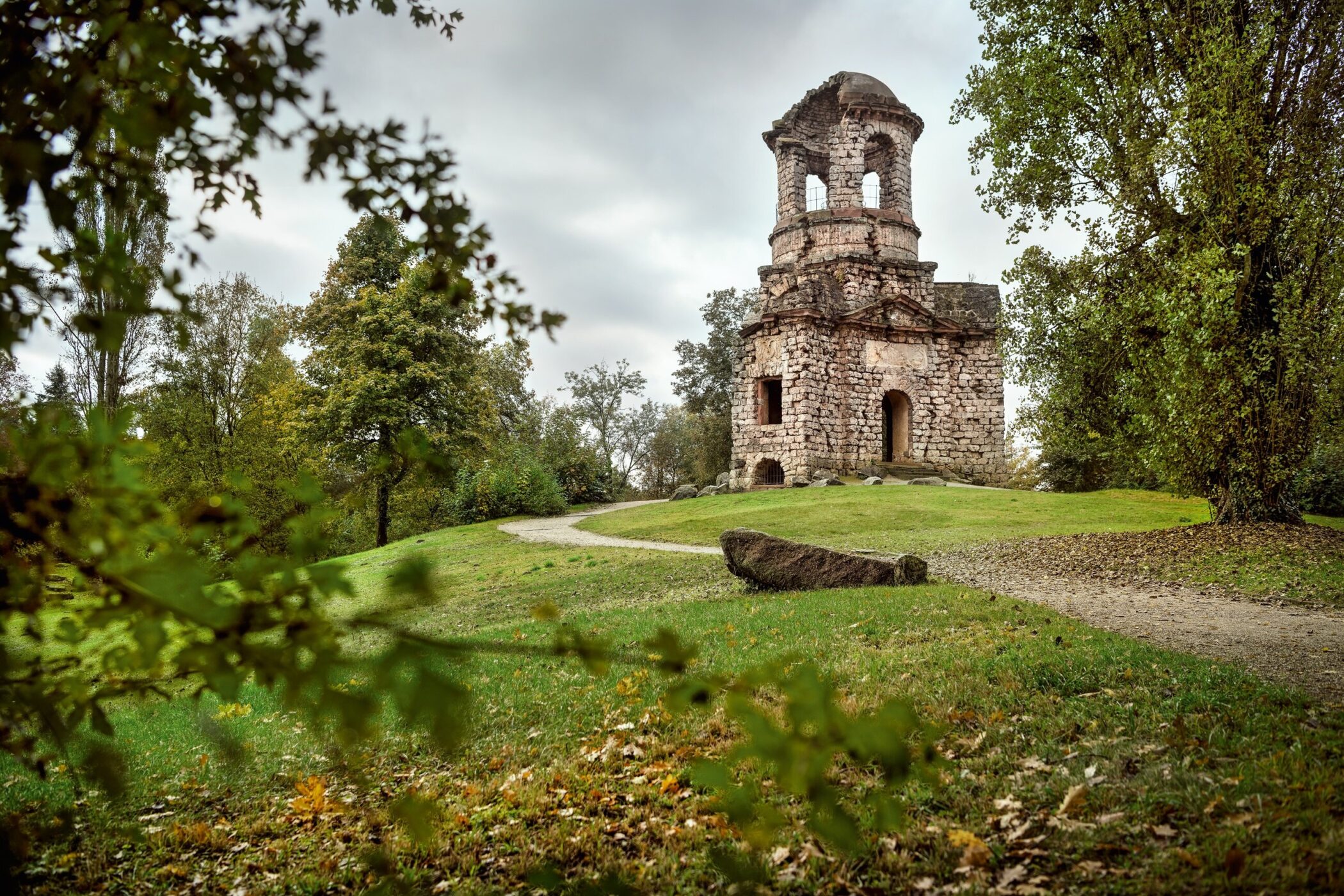 Steinruine mit Turm auf einer Wiese, umgeben von Bäumen und einem Weg.