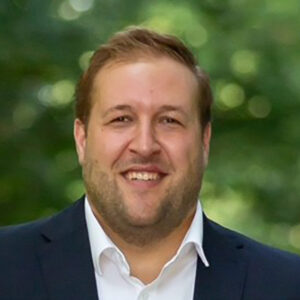 Ein lächelnder Mann in Anzug vor unscharfem, grünem Hintergrund.