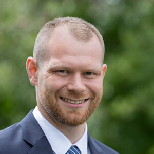 Ein lächelnder Mann im Anzug vor unscharfem, grünem Hintergrund.