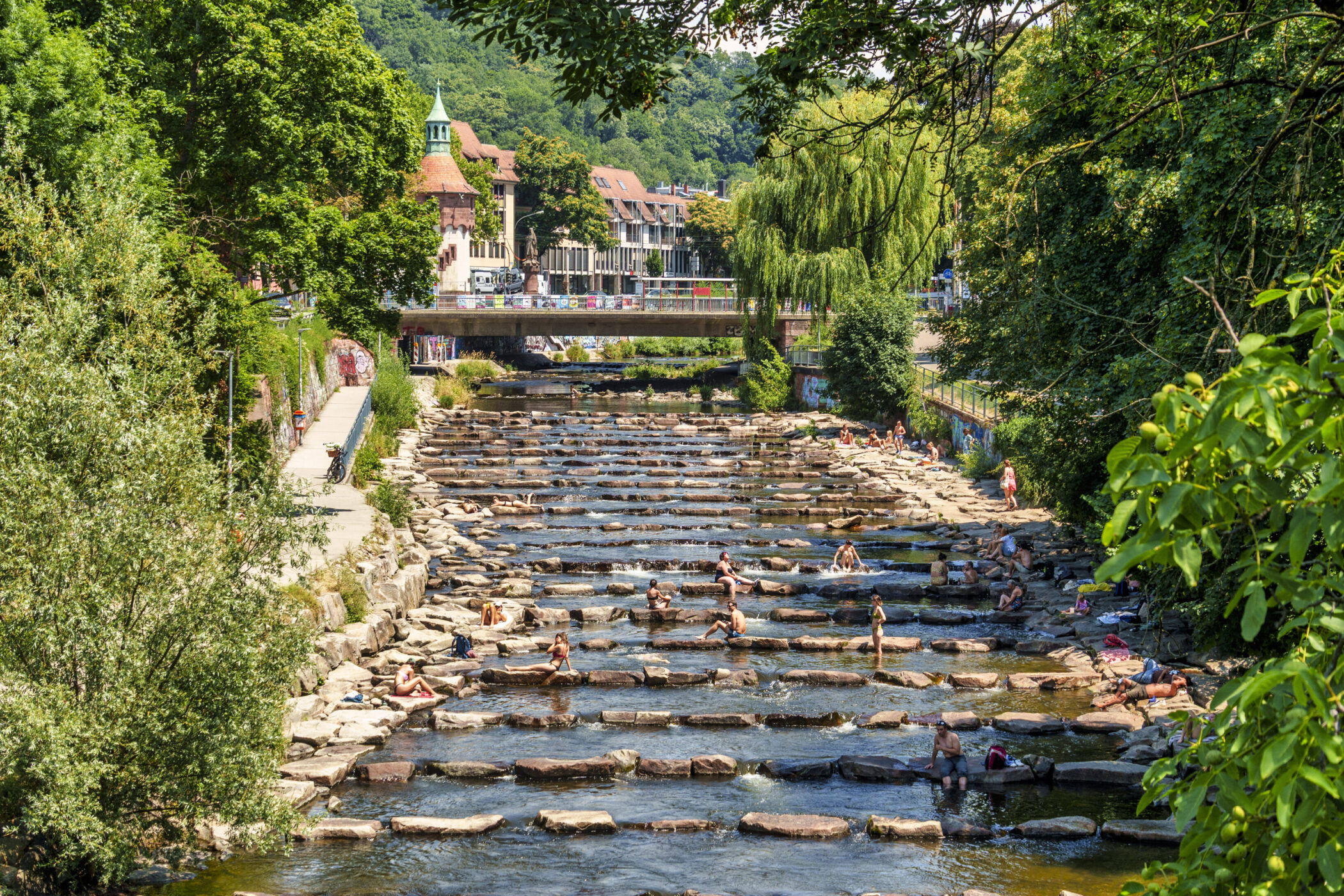 Fluss mit Steinen und Menschen, die im Wasser sitzen; Brücke und Gebäude im Hintergrund.