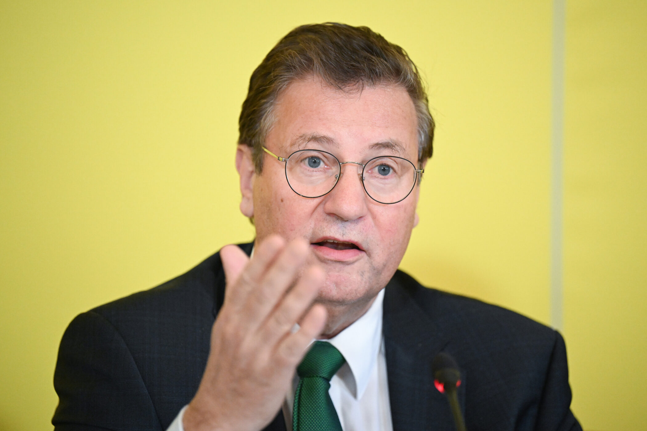 Mann mit Brille spricht, gelber Hintergrund, Mikrofon sichtbar.