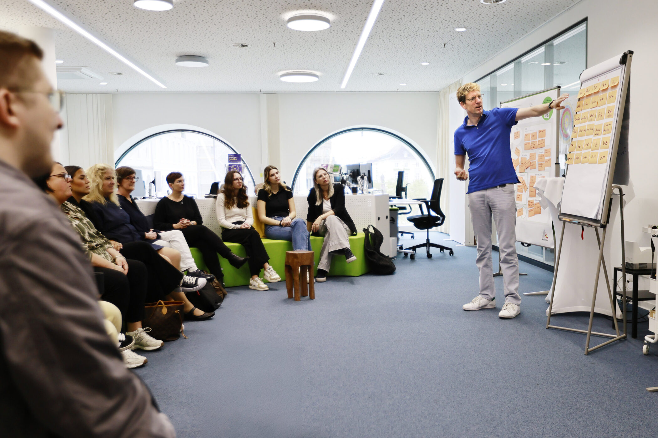 Person zeigt auf Flipchart, Gruppe von neun Personen sitzt und hört zu.