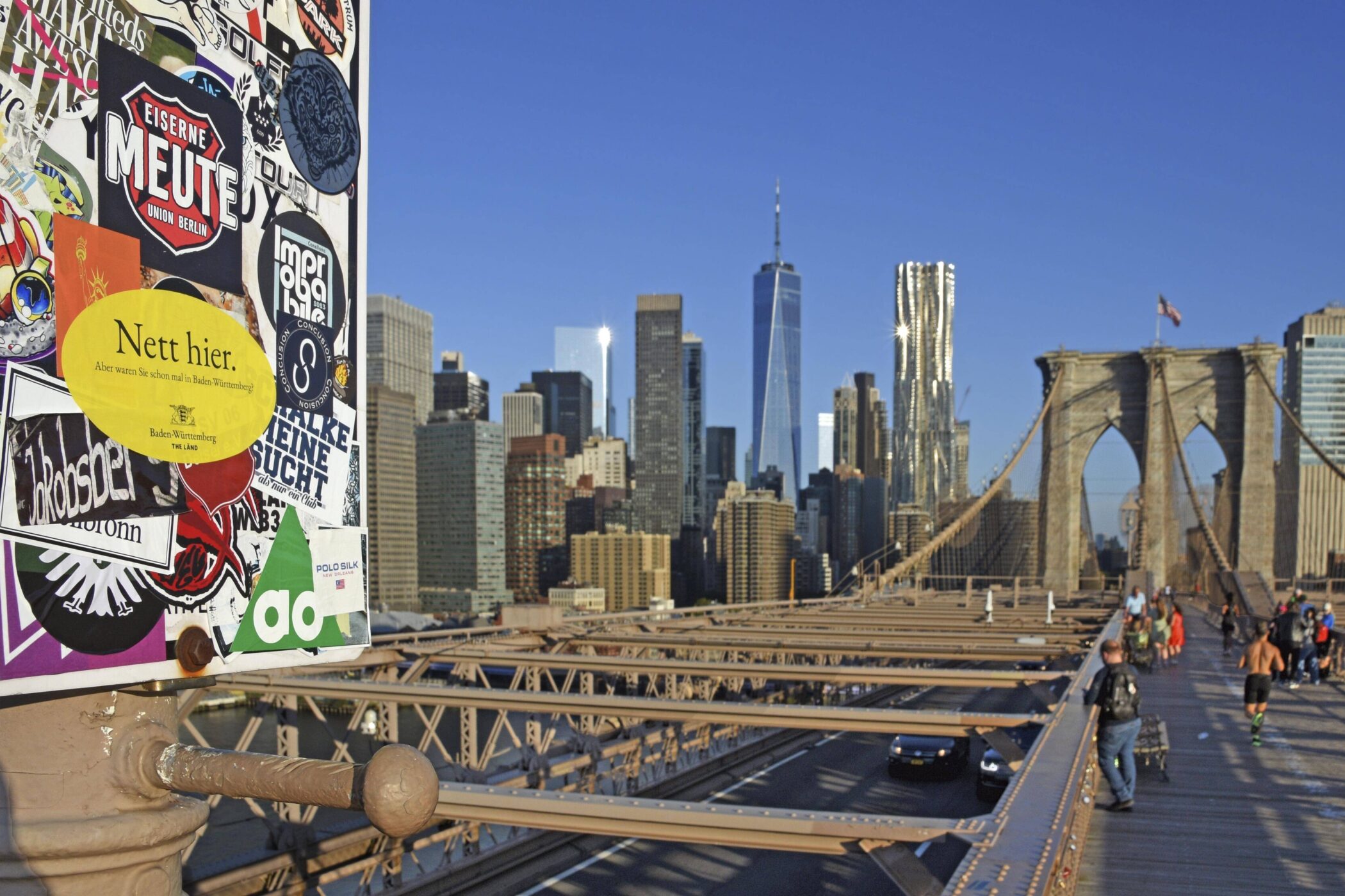 Aufgeklebte Sticker, Skyline und Brücke im Hintergrund, Menschen auf Gehweg.