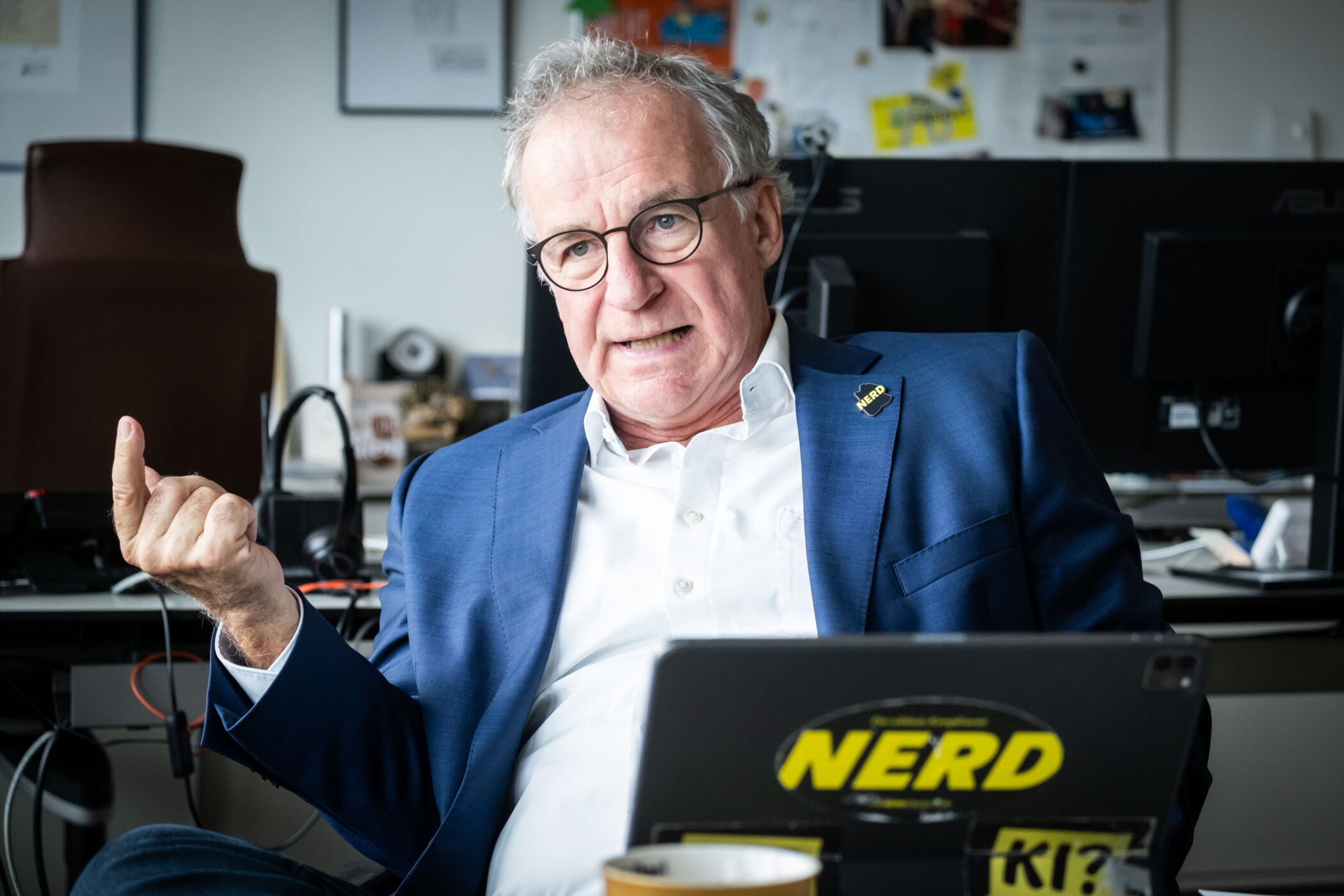 Älterer Mann im Anzug gestikuliert vor einem Laptop mit "NERD"-Aufkleber.