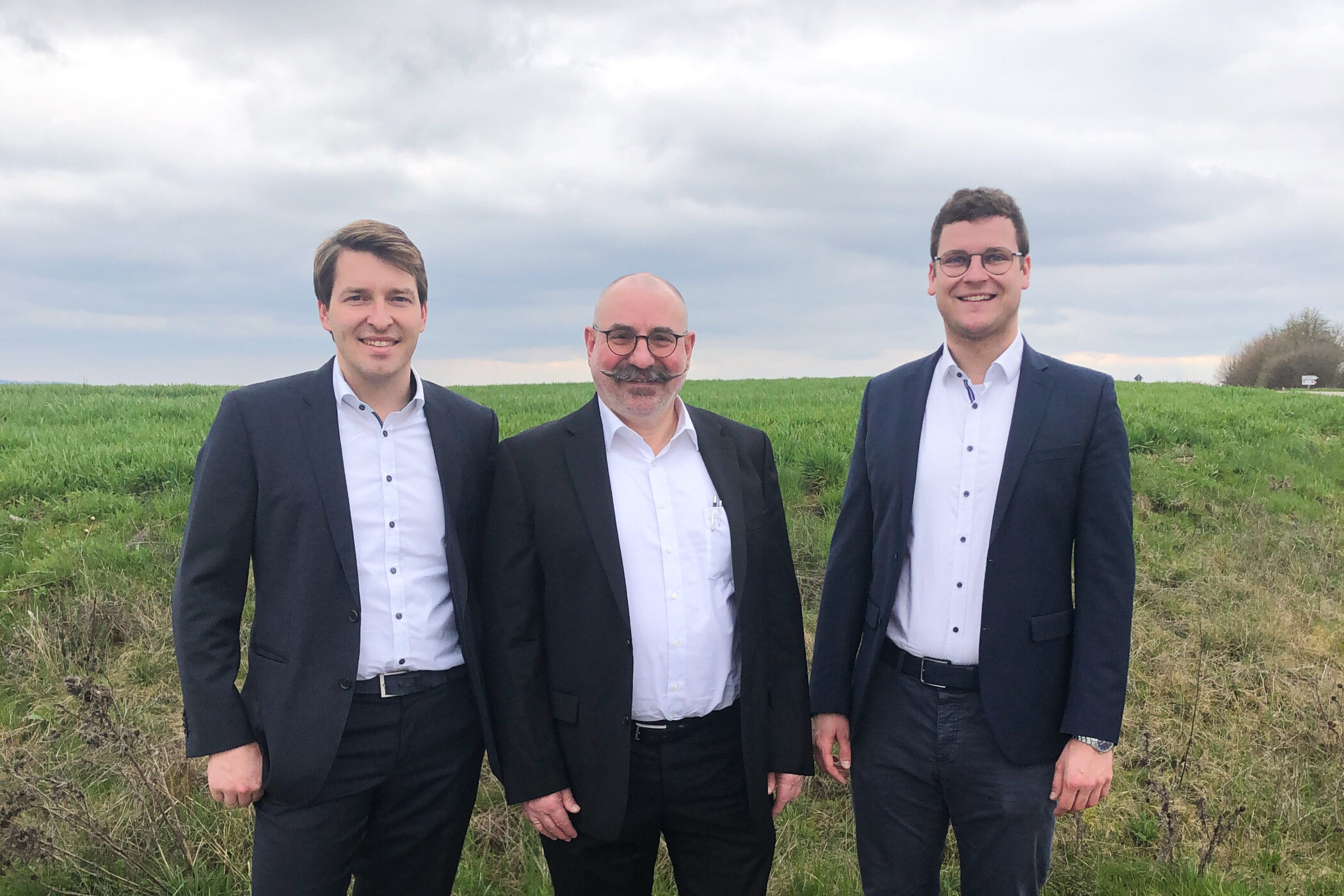 Drei Männer in Anzügen stehen auf einer Wiese, bewölkter Himmel.