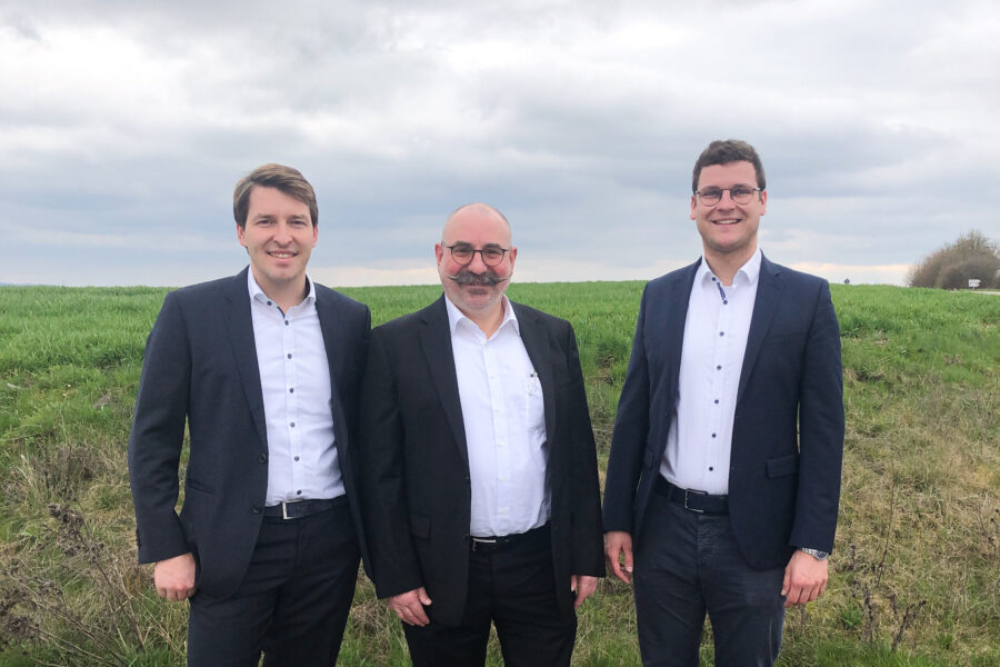 Drei Männer in Anzügen stehen auf einer Wiese, bewölkter Himmel.