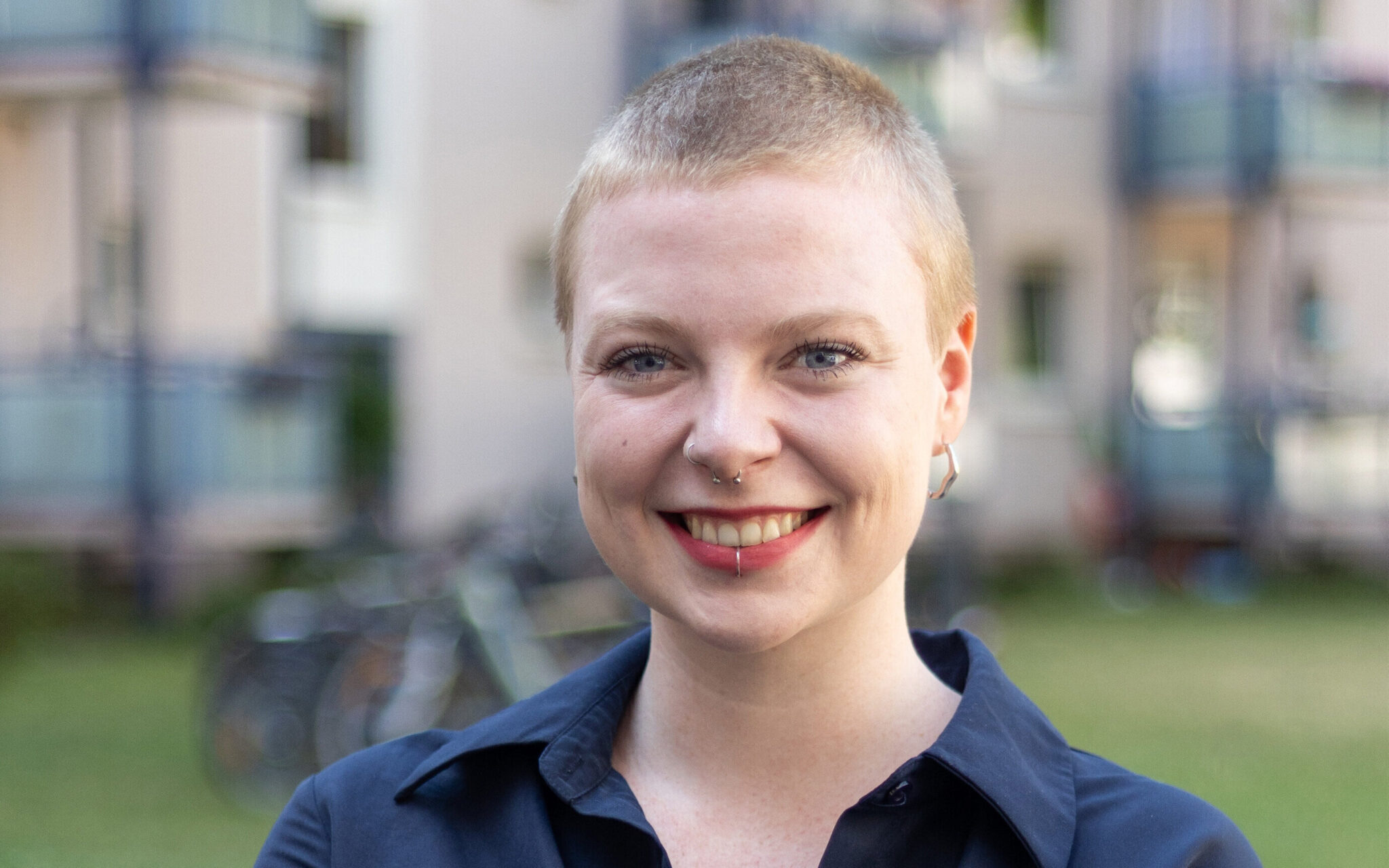 Eine Person mit kurzem Haar und Piercings lächelt im Freien.