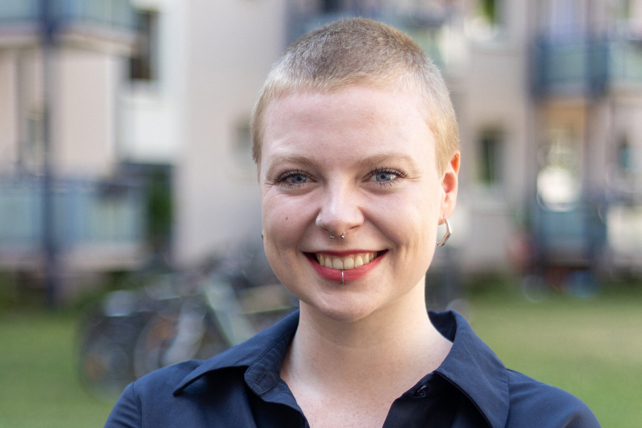 Eine Person mit kurzem Haar und Piercings lächelt im Freien.